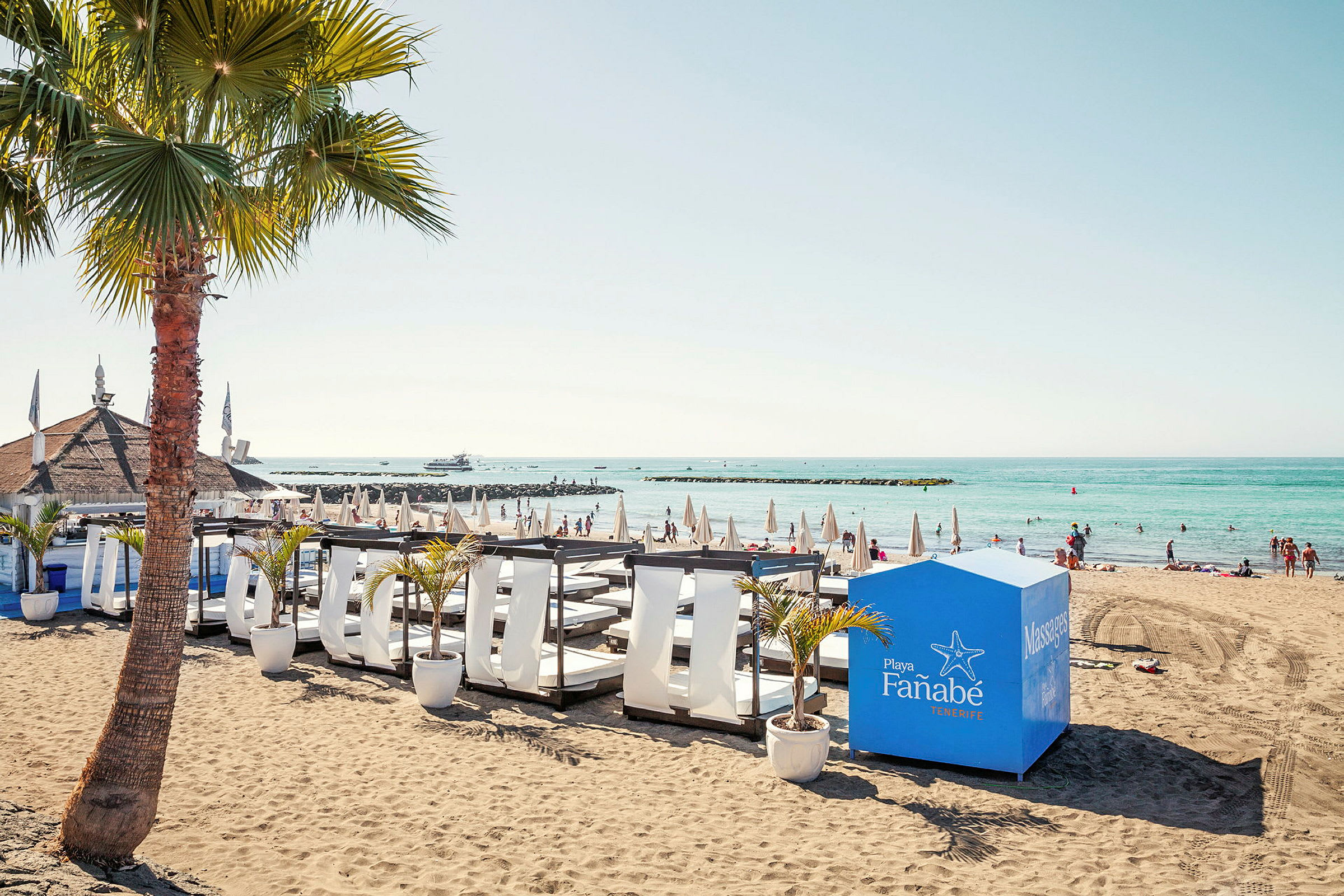 En strand med solstolar, parasoller, en palm och en massagekiosk vid havet. (AI-genererad text)