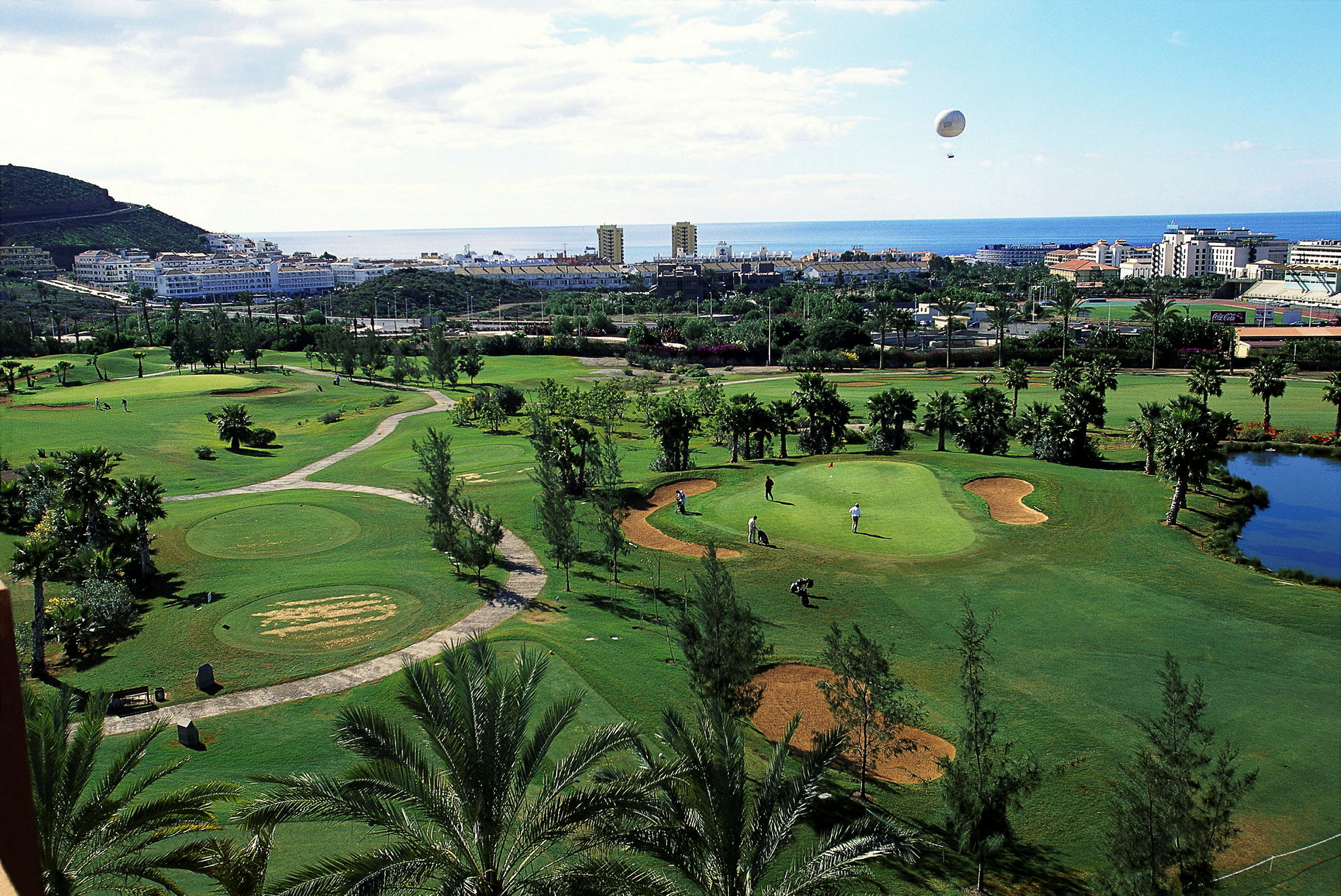 Golfbana med palmer, bunkrar och gröna fairways, med byggnader och havet i bakgrunden. (AI-genererad text)