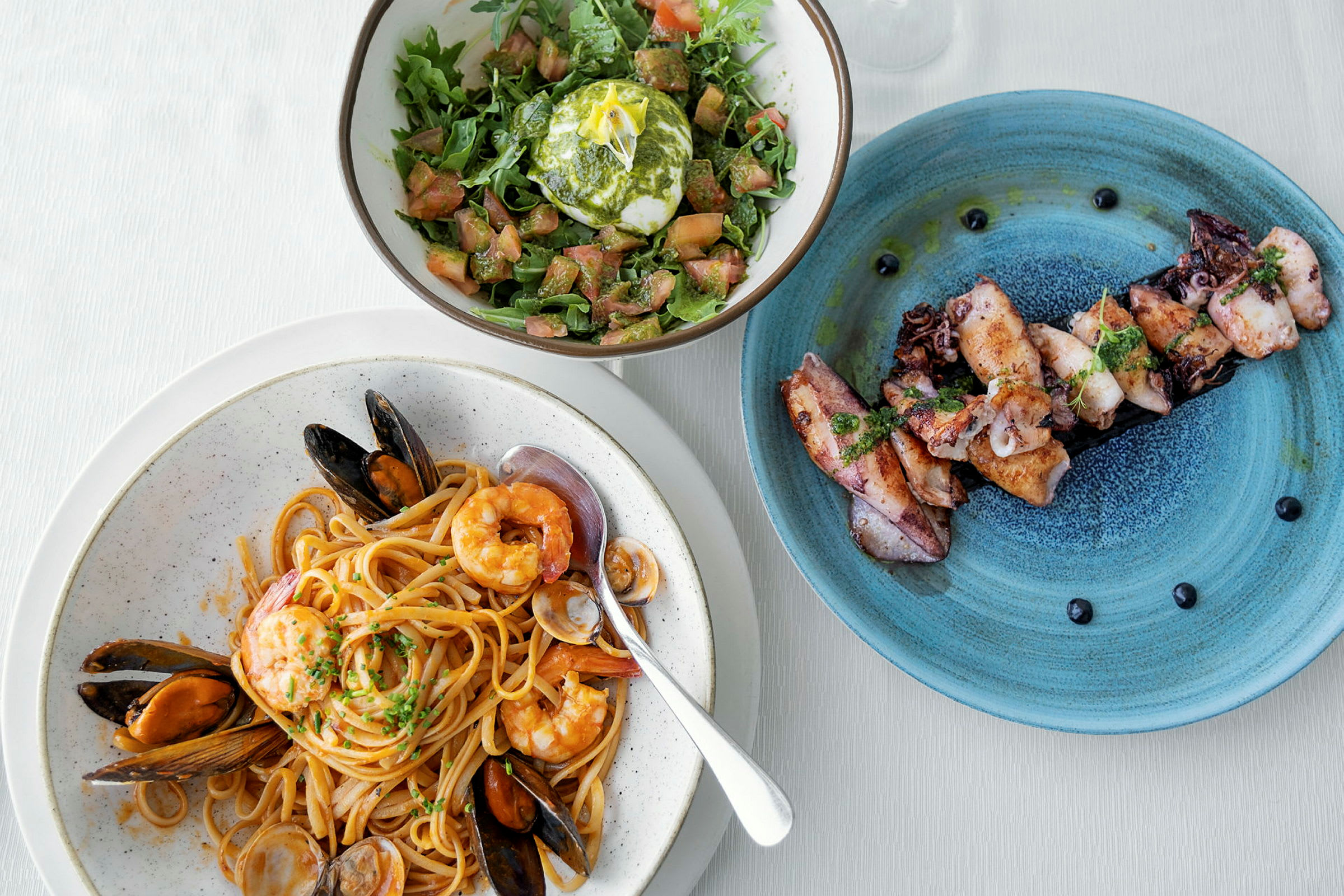 En tallrik med pasta och skaldjur, en sallad med mozzarella, och en tallrik med grillad bläckfisk. (AI-genererad text)