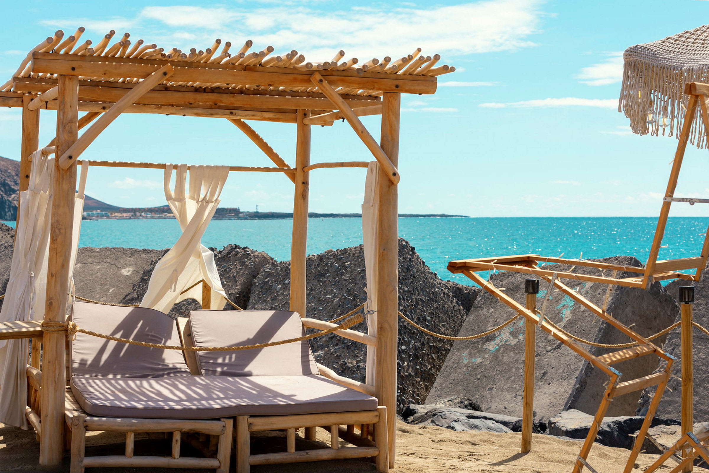 Två solstolar under ett träpergola på en sandstrand med utsikt över havet och stora stenblock. (AI-genererad text)