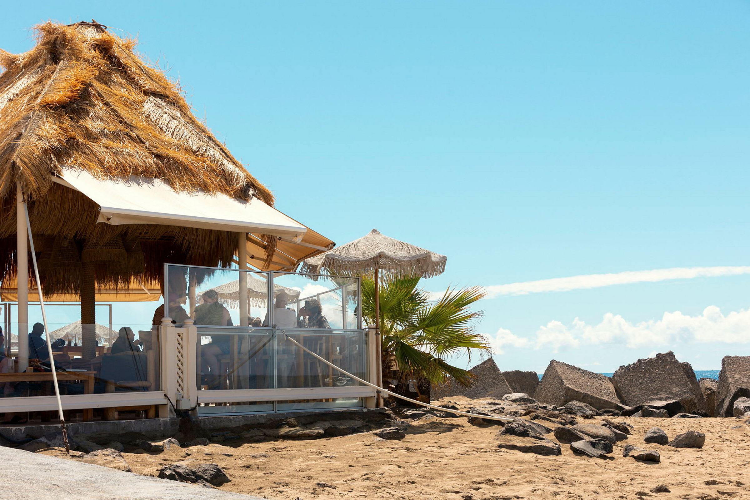 En strandbar med tak av halm och parasoller, flankerad av sand och klippor. (AI-genererad text)