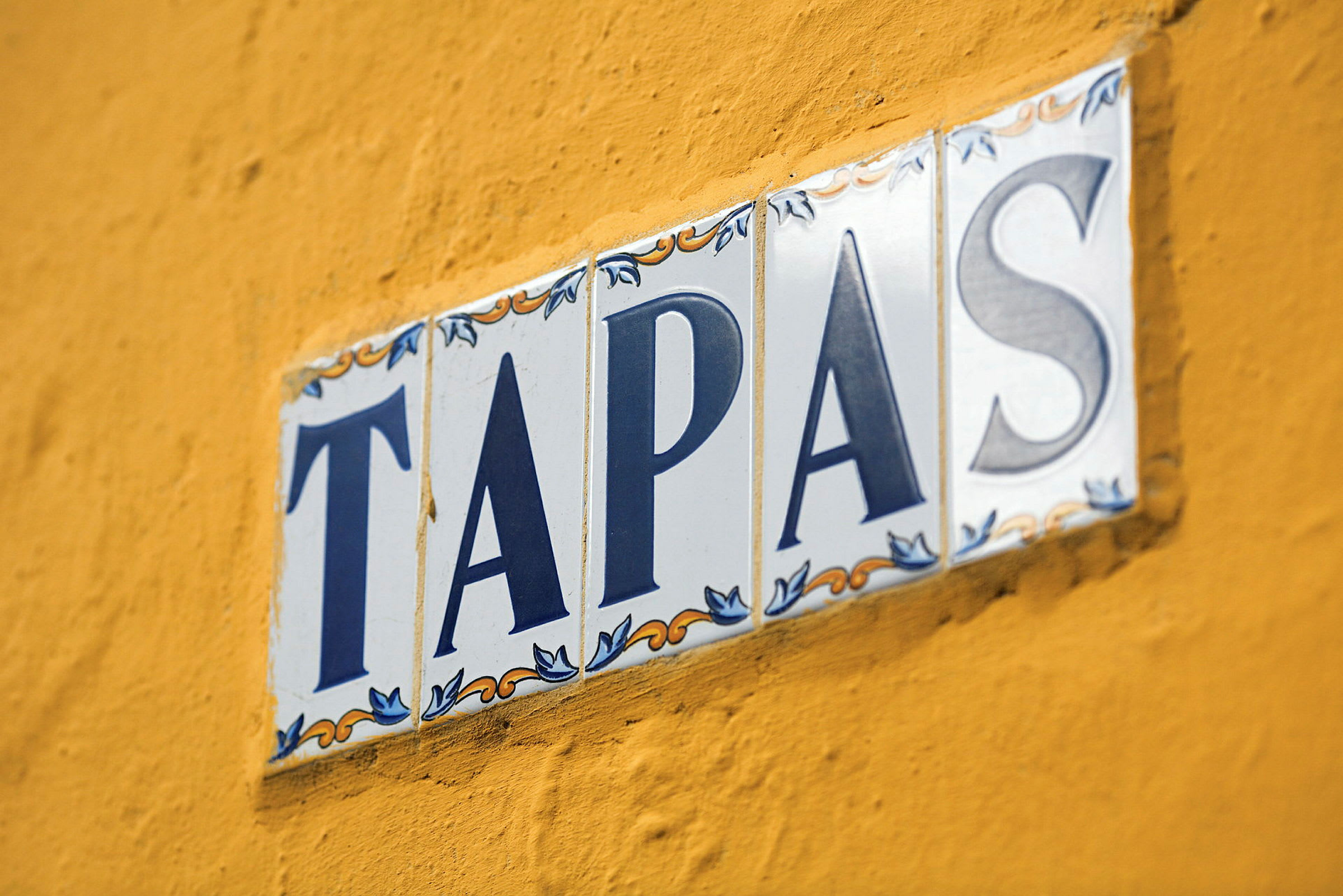 En skylt med texten 'TAPAS' på vita keramiska plattor mot en gul vägg. (AI-genererad text)
