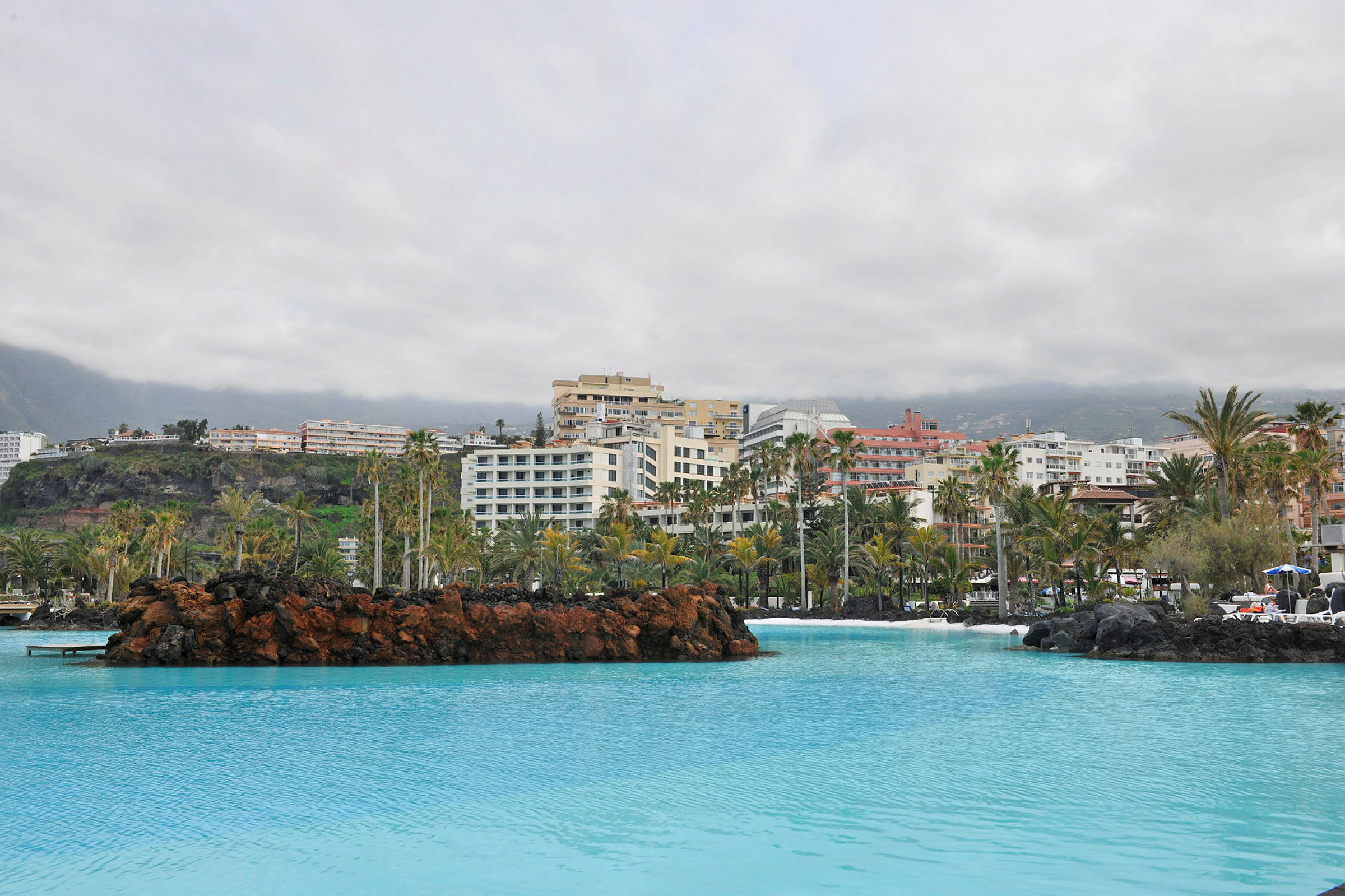 Hotellbyggnader bakom en pool med palmer och lavaformationer. (AI-genererad text)