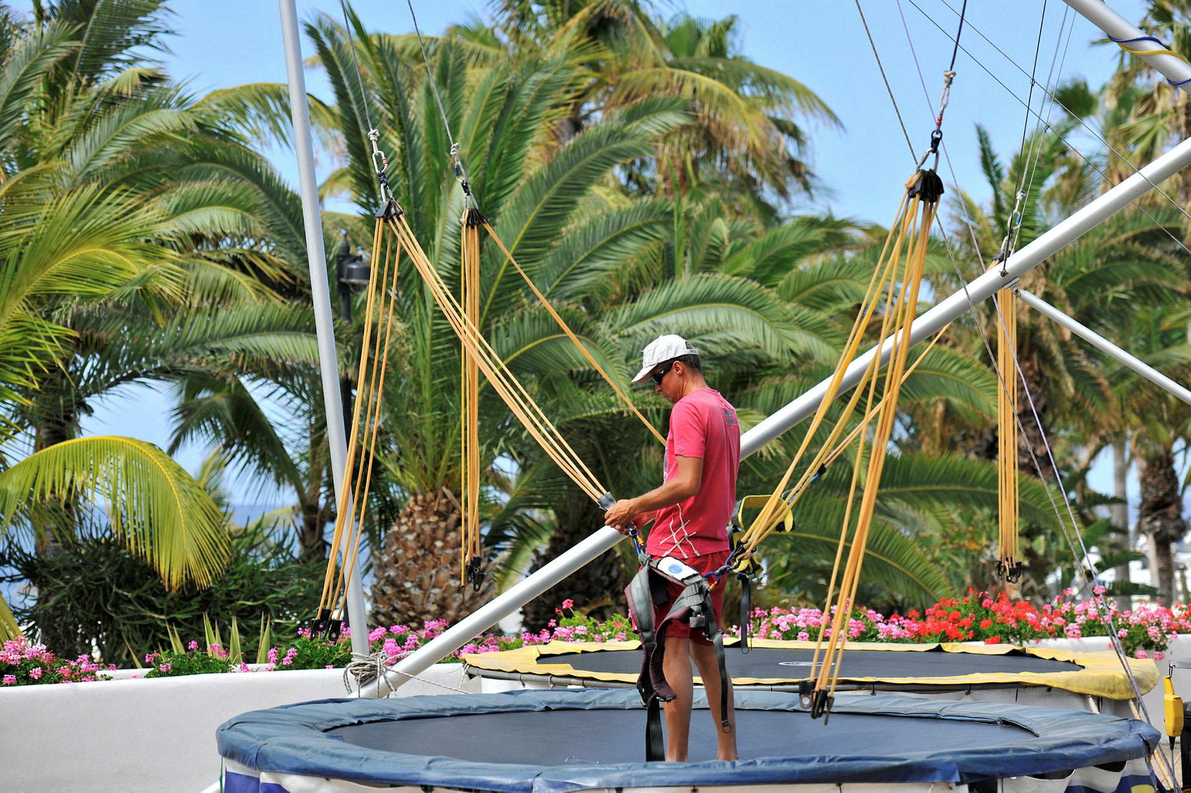 En person står vid en trampolin med sele och linor. Bakgrunden är fylld med grönskande palmträd. (AI-genererad text)