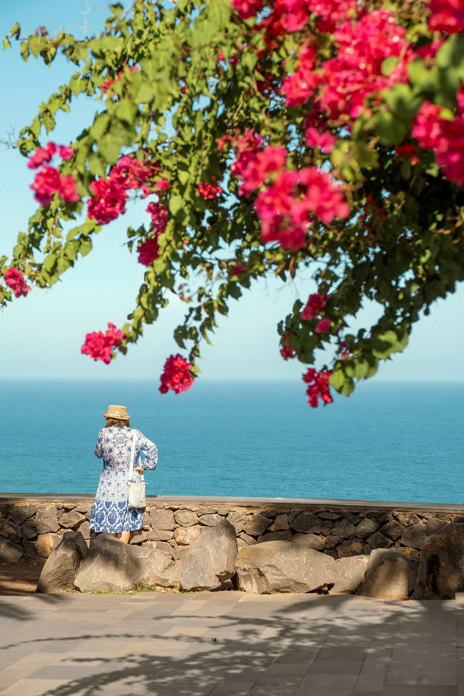 Person som står vid en stenmur med utsikt över havet under en blommande bougainvillea. (AI-genererad text)