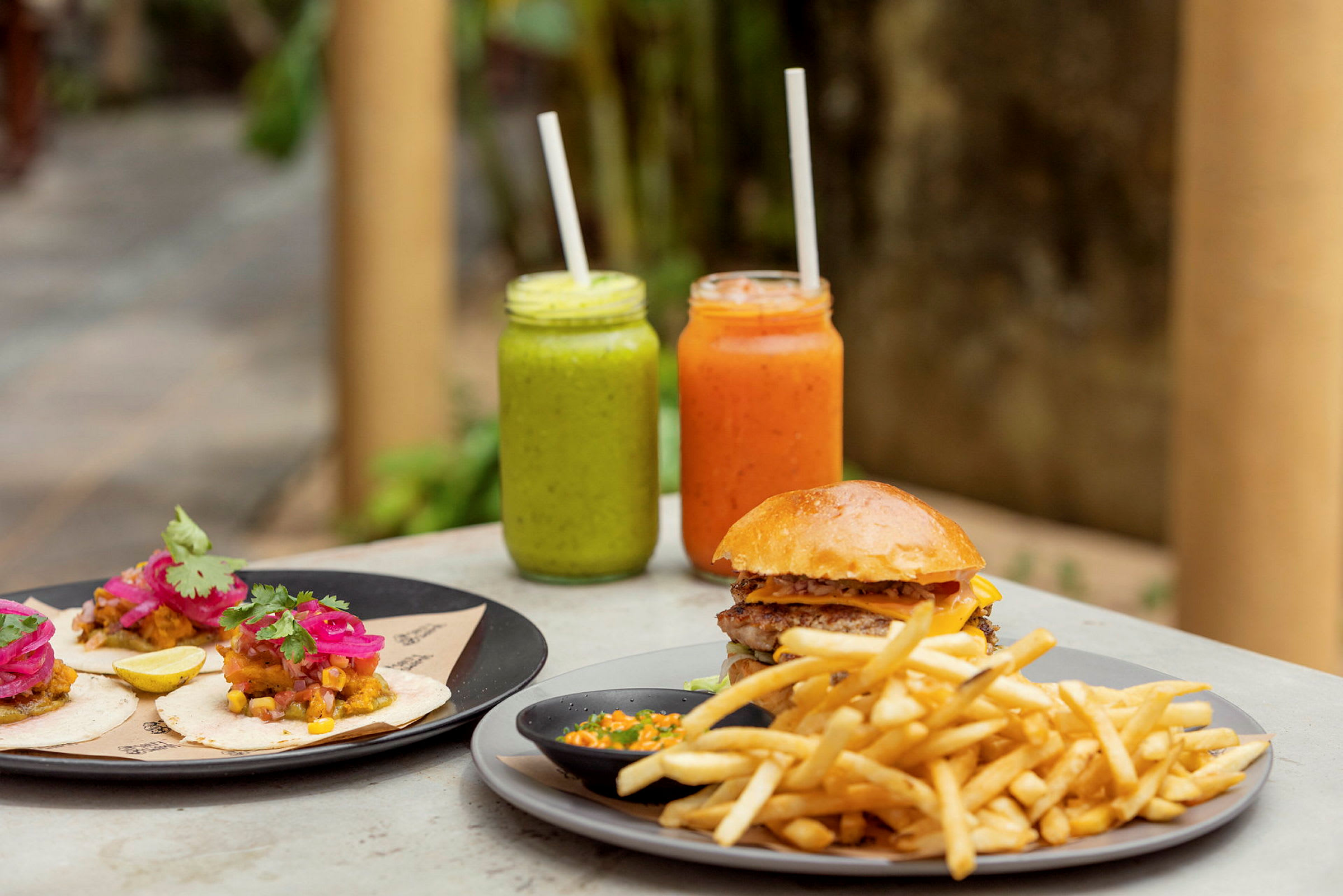 På bordet står en tallrik med tacos, en med hamburgare och pommes frites samt två glas med färgglada drycker. (AI-genererad text)