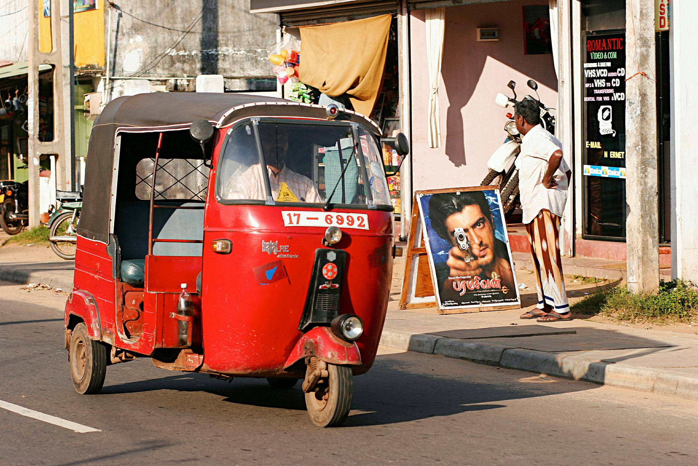 En röd tuk-tuk kör förbi en butik på en solig gata. (AI-genererad text)