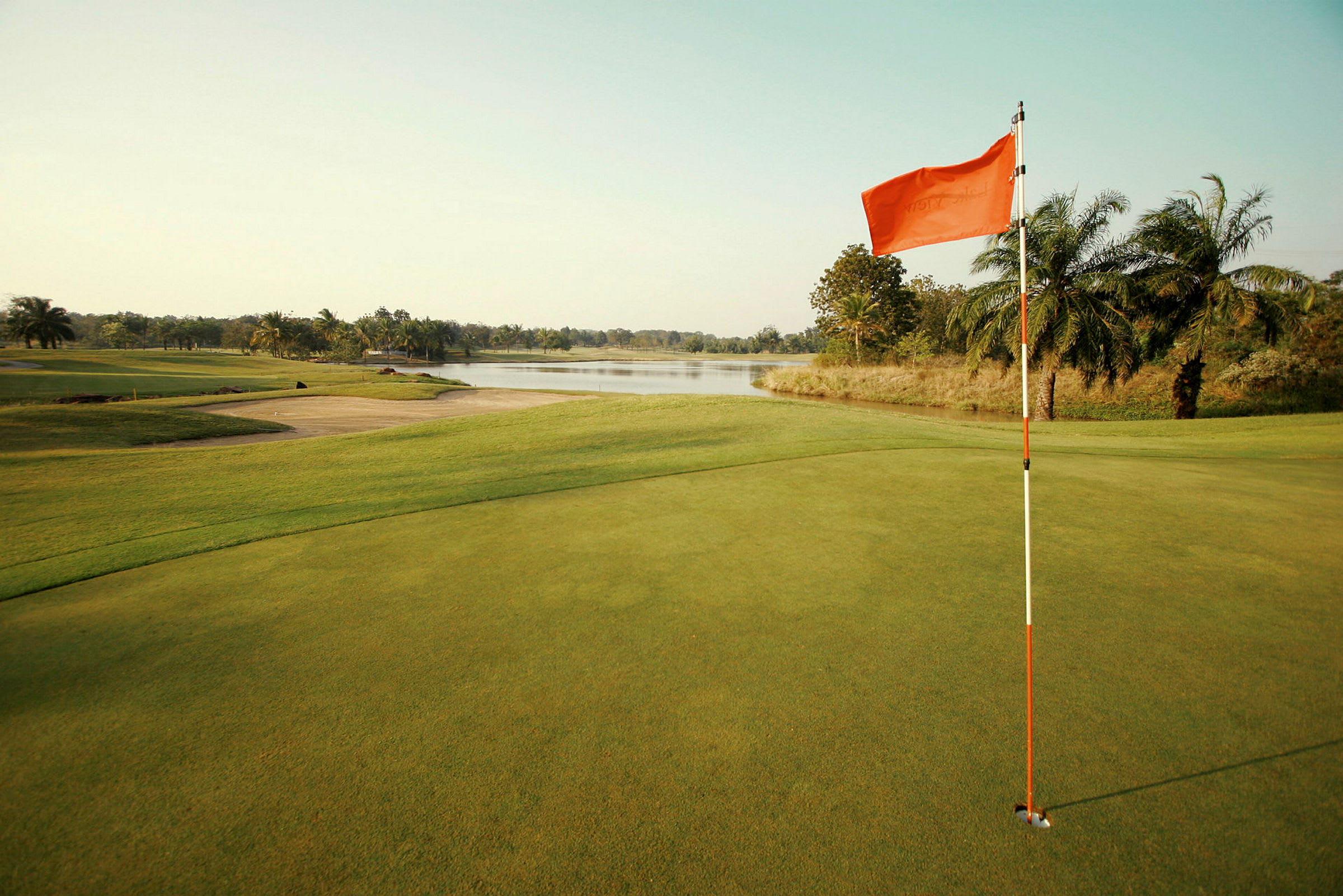 Golfbana med flagga vid green nära en sjö och omgiven av palmer. (AI-genererad text)