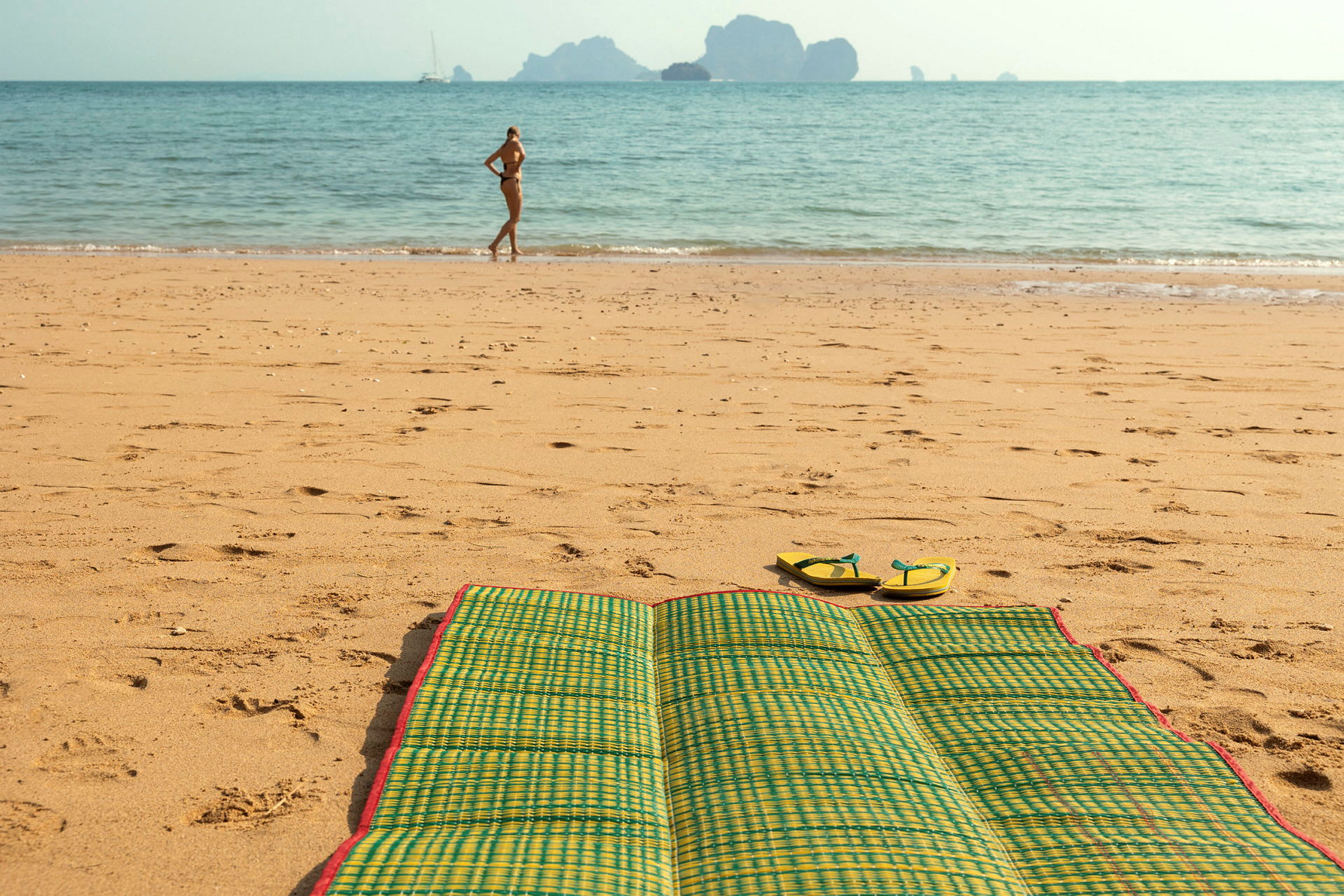 En strand med en grön strandmatta och ett par flip-flops i förgrunden. En person står vid vattenkanten i bakgrunden. (AI-genererad text)