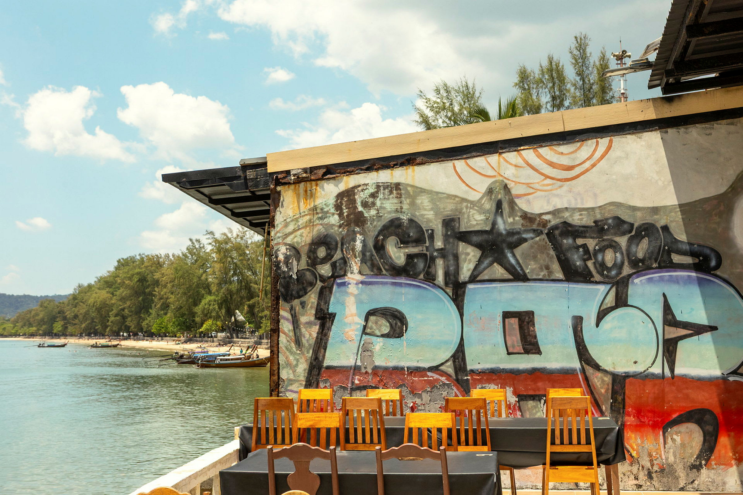 En utsikt över en strand med vatten, träd och båtar. Framför bilden syns en vägg med graffiti, samt stolar och bord. (AI-genererad text)