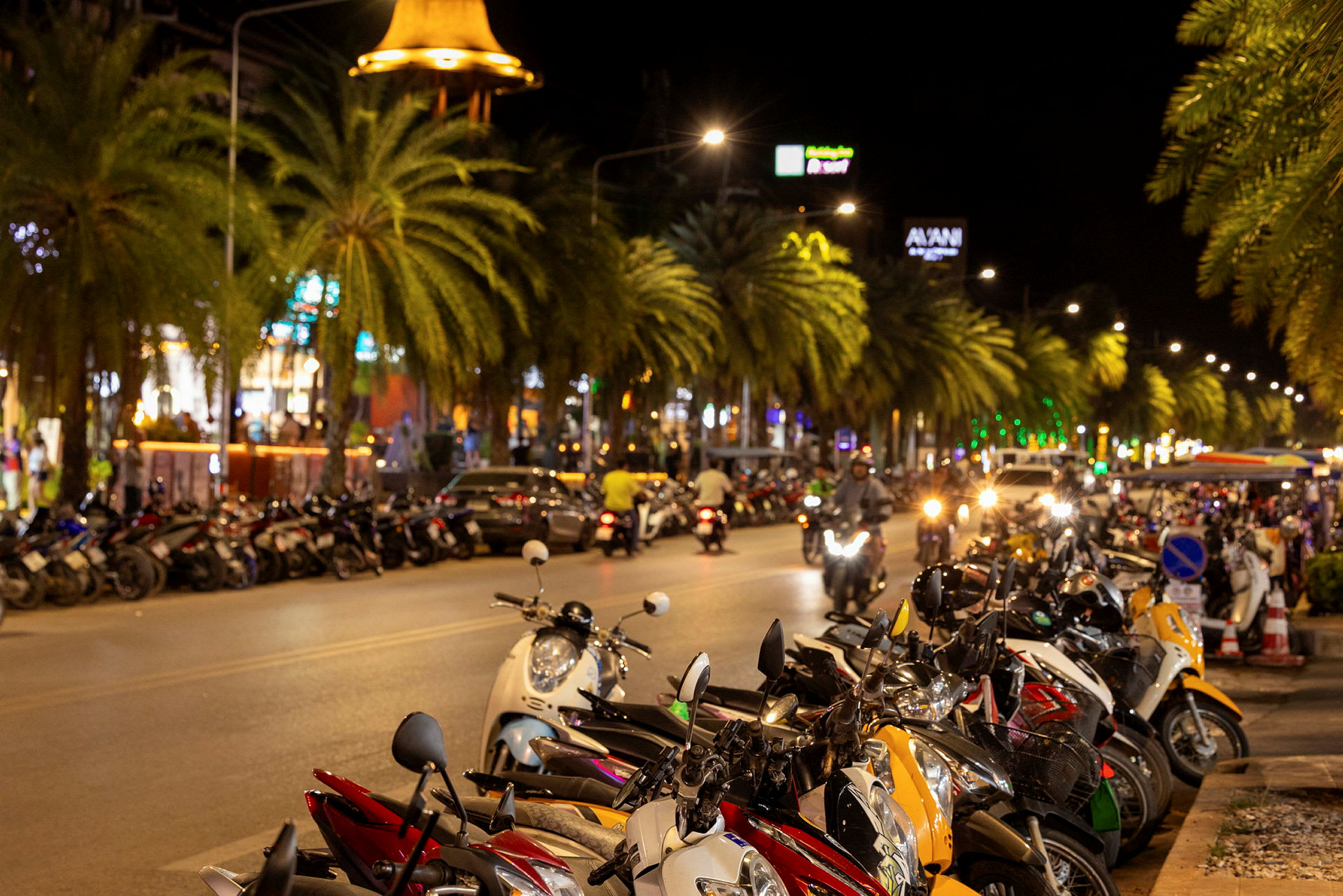 Motorcyklar parkerade längs en livlig gata kantad av palmer, med kvällsbelysning och trafikrörelse. (AI-genererad text)