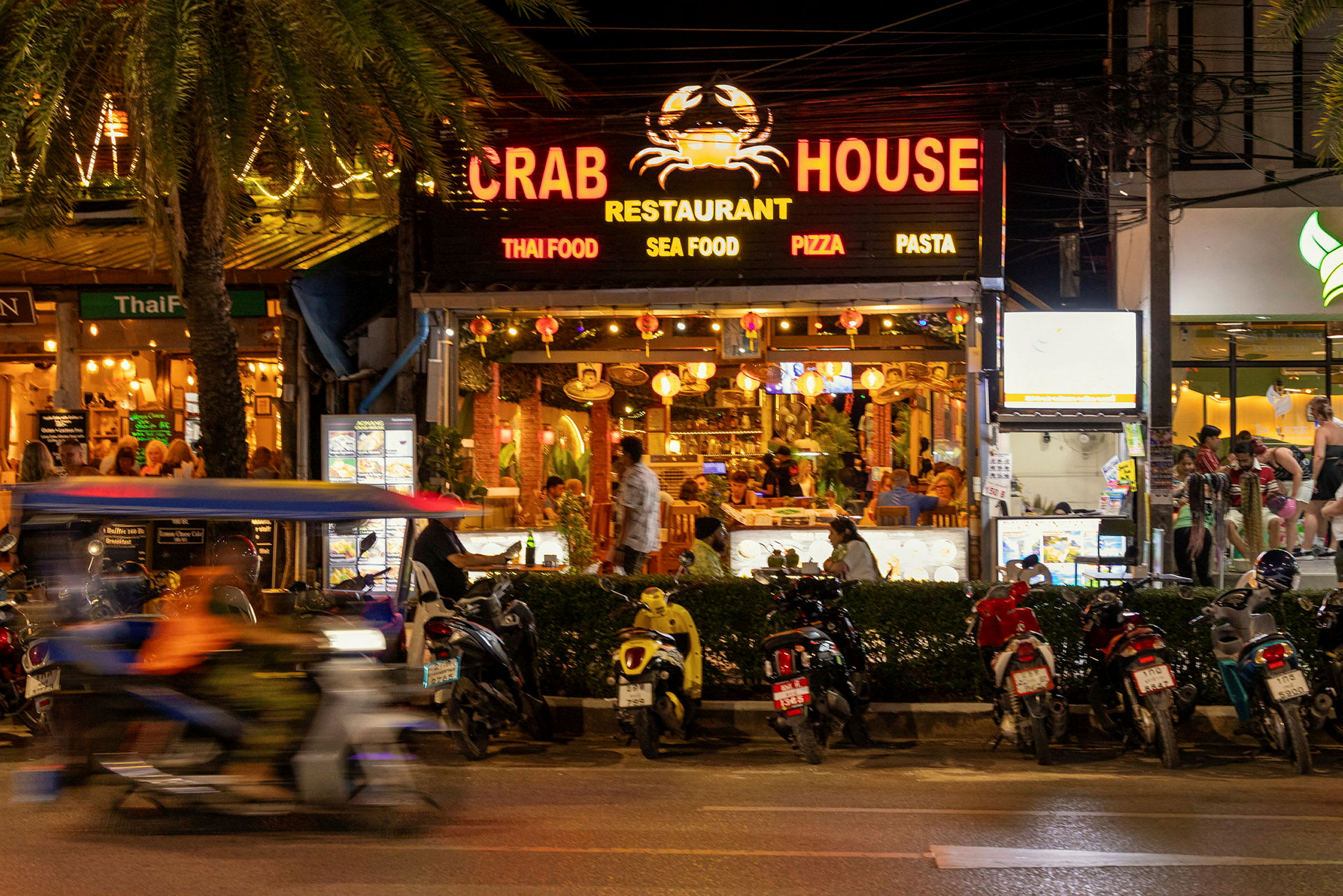 Bild av en livlig gata med restaurangen Crab House i fokus. Flera motorcyklar står parkerade framför restaurangen och en motorcykel med en passagerare kör förbi. (AI-genererad text)