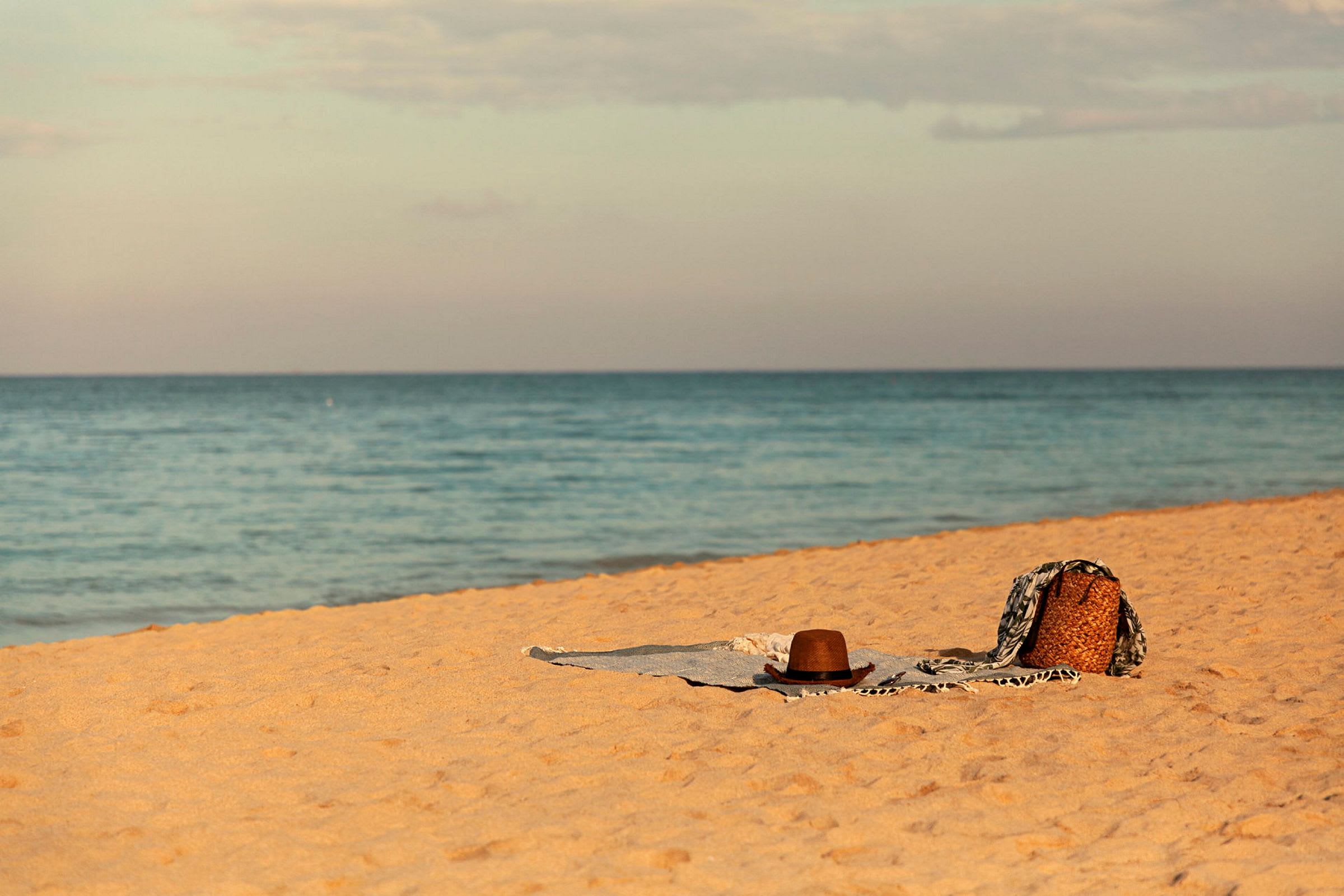 En strand med gul sand där ett hatt, en korg och en filt ligger utbredda. Havet är synligt i bakgrunden. (AI-genererad text)