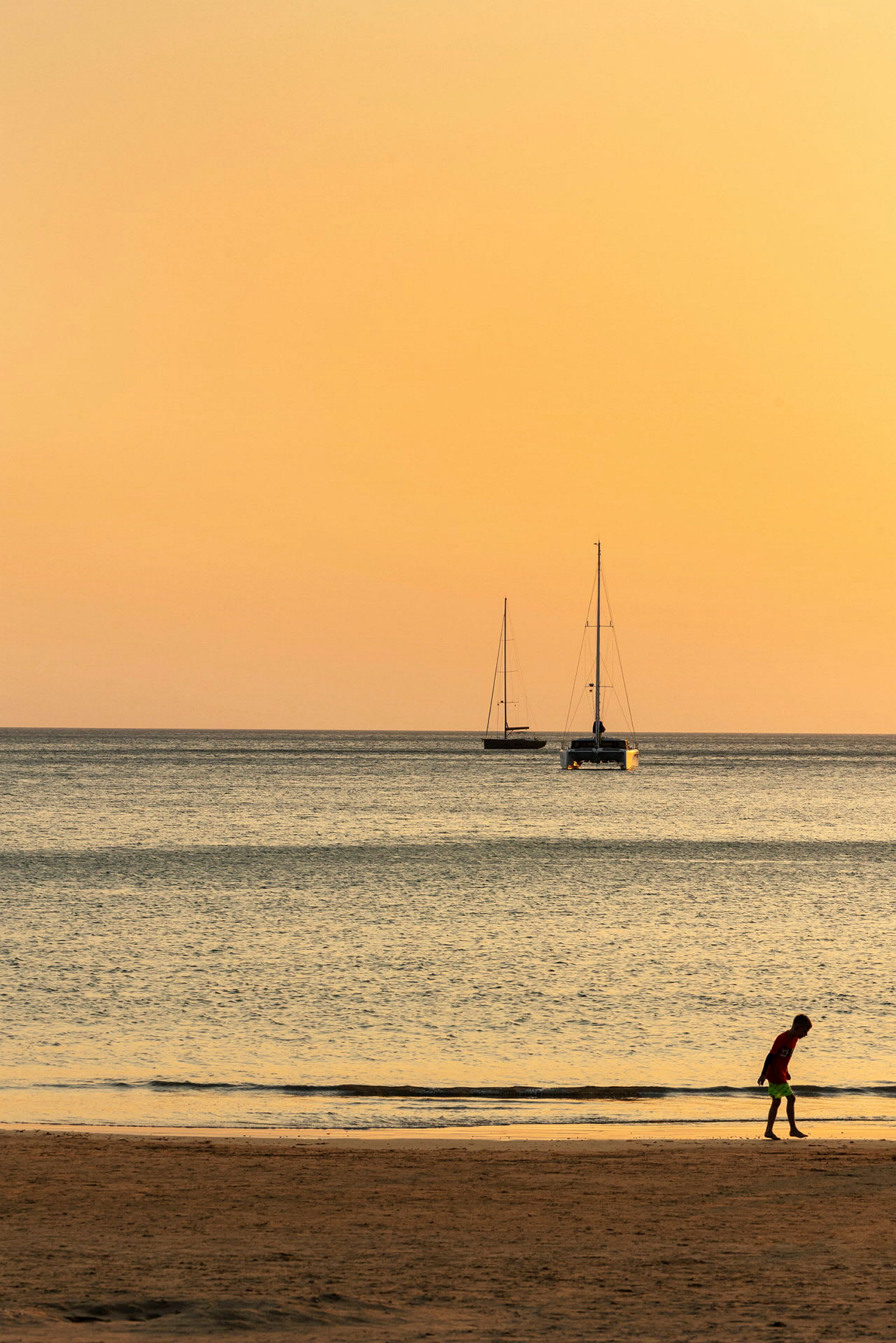 En strand vid havet under en orange solnedgång med två segelbåtar i bakgrunden och en person som promenerar i sanden. (AI-genererad text)