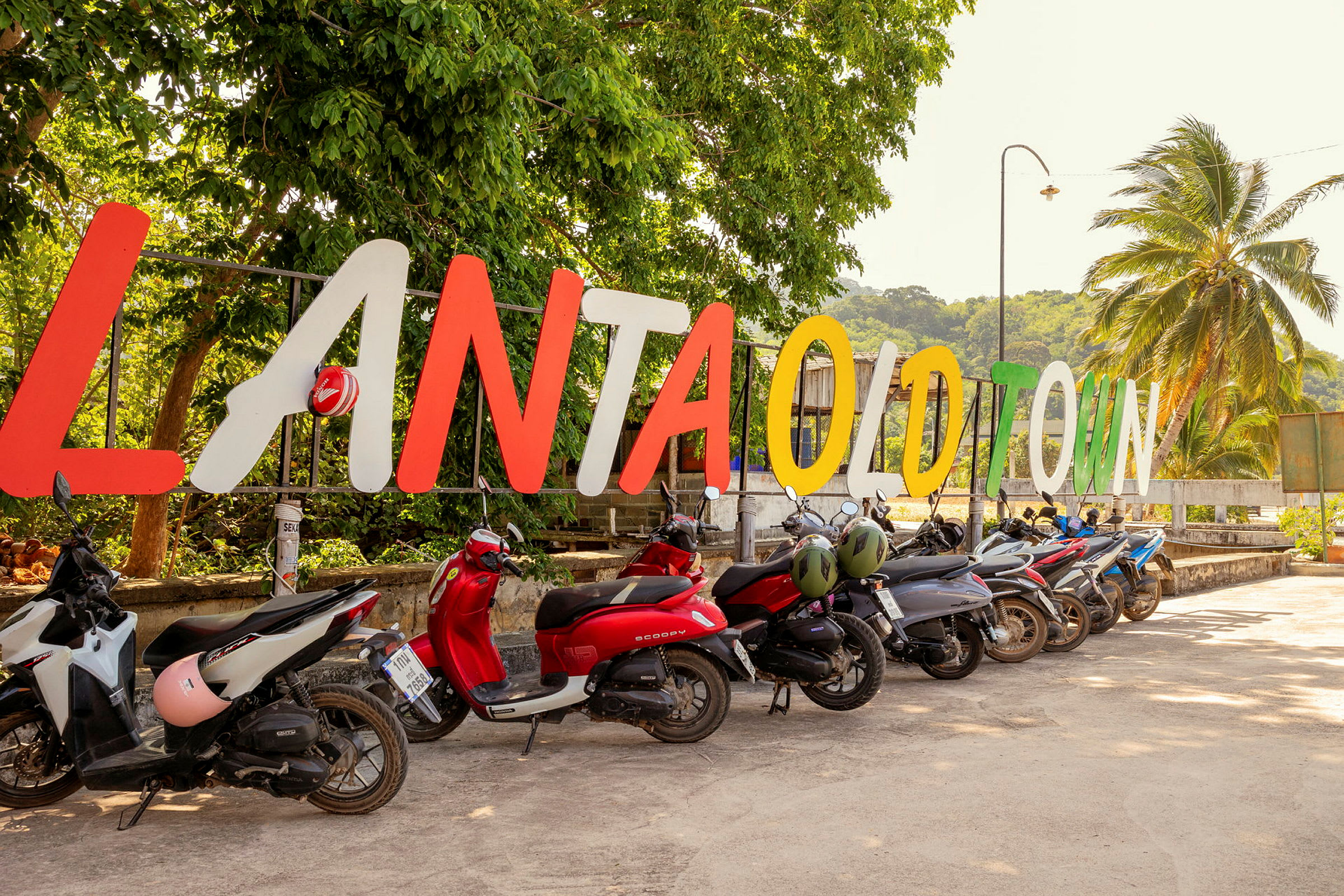 En uppställd skylt med texten 'Lanta Old Town' bakom en rad parkerade motorcyklar. I bakgrunden finns grönska och en palm. (AI-genererad text)