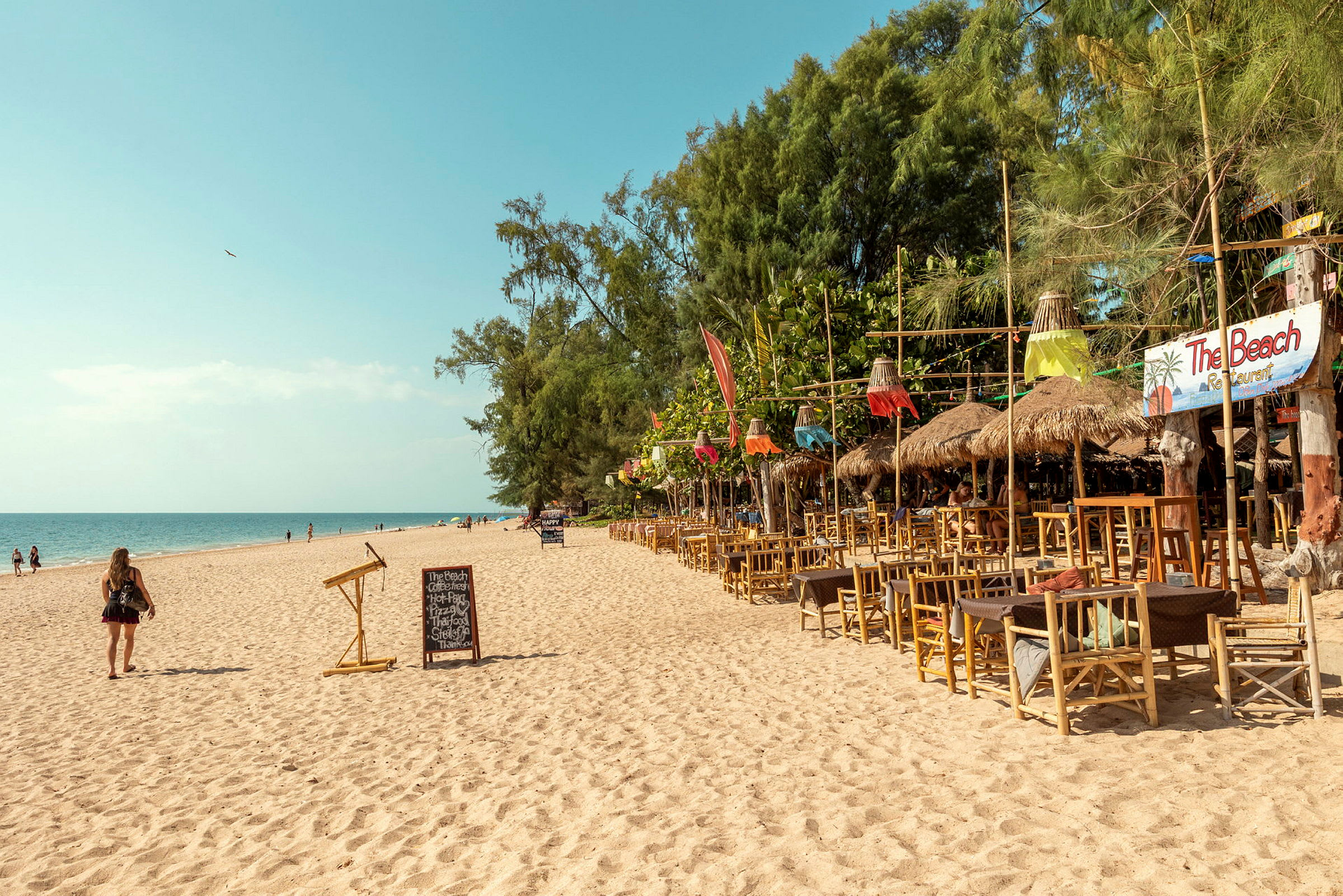 En sandstrand med en rad trämöblerade restaurangområden med dekorationer till höger och människor som promenerar vid havet. (AI-genererad text)