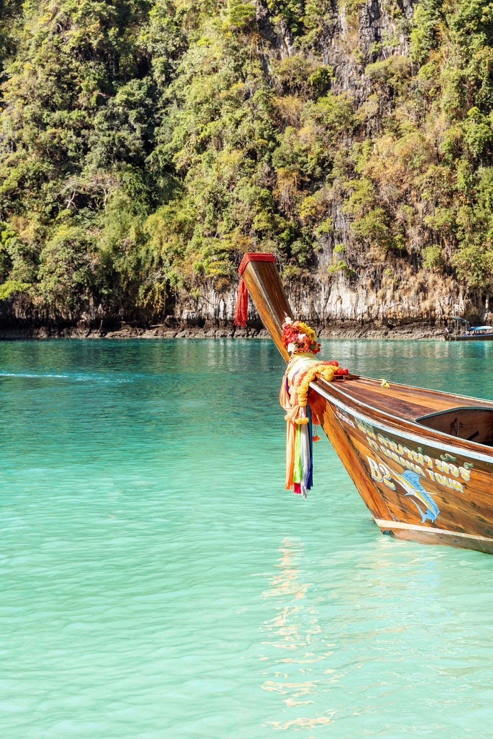 En traditionell thailändsk båt prydd med färgglada band och dekorationer flyter på kristallklart vatten, med en grönskande klippa i bakgrunden. (AI-genererad text)