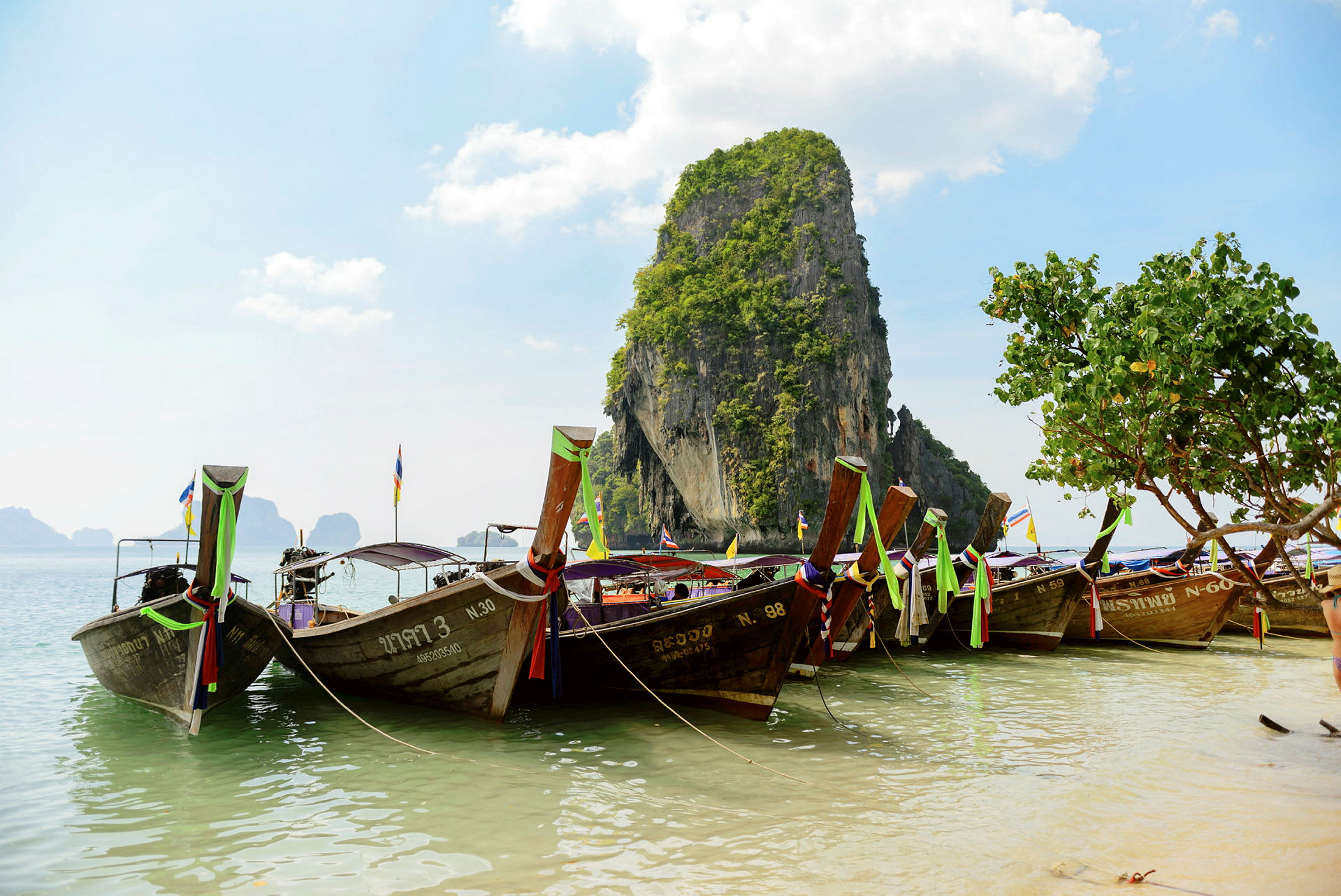 Traditionella thailändska båtar förtöjda vid en strand med en kalkstensklippa i bakgrunden. (AI-genererad text)