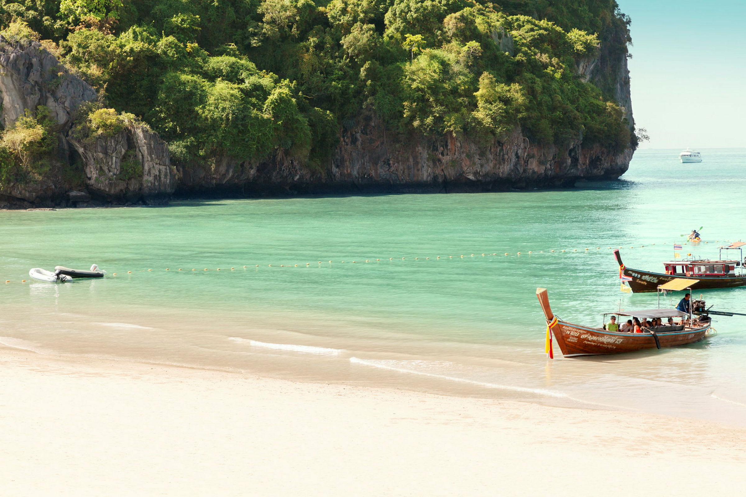 En sandstrand med klart grönt vatten, båtar och grönskande klippor i bakgrunden. (AI-genererad text)