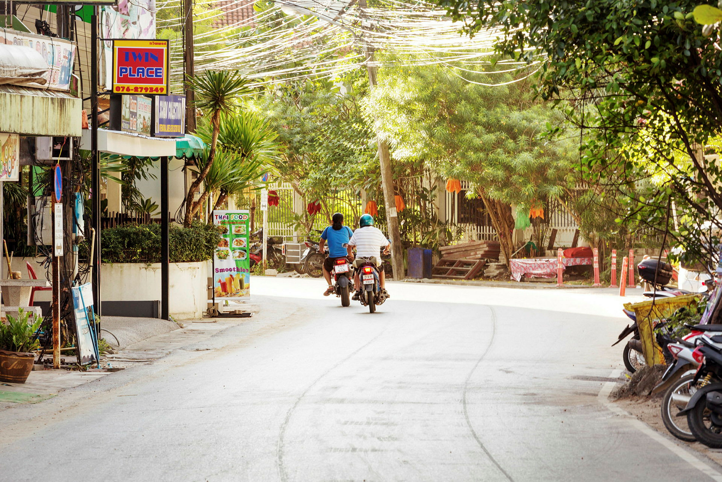 Två personer på motorcyklar kör på en väg omgiven av grönska och byggnader. (AI-genererad text)