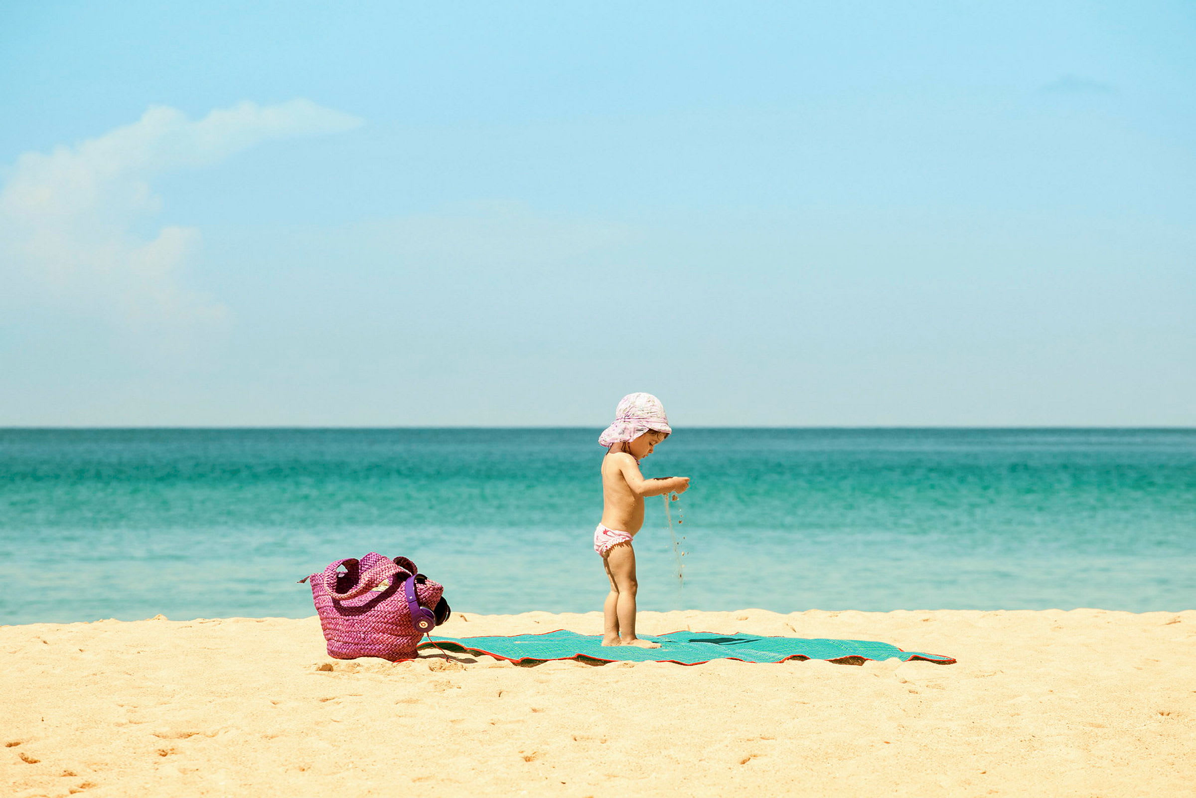 Ett barn på en strand med ett hav i bakgrunden och en lila väska på sanden. (AI-genererad text)