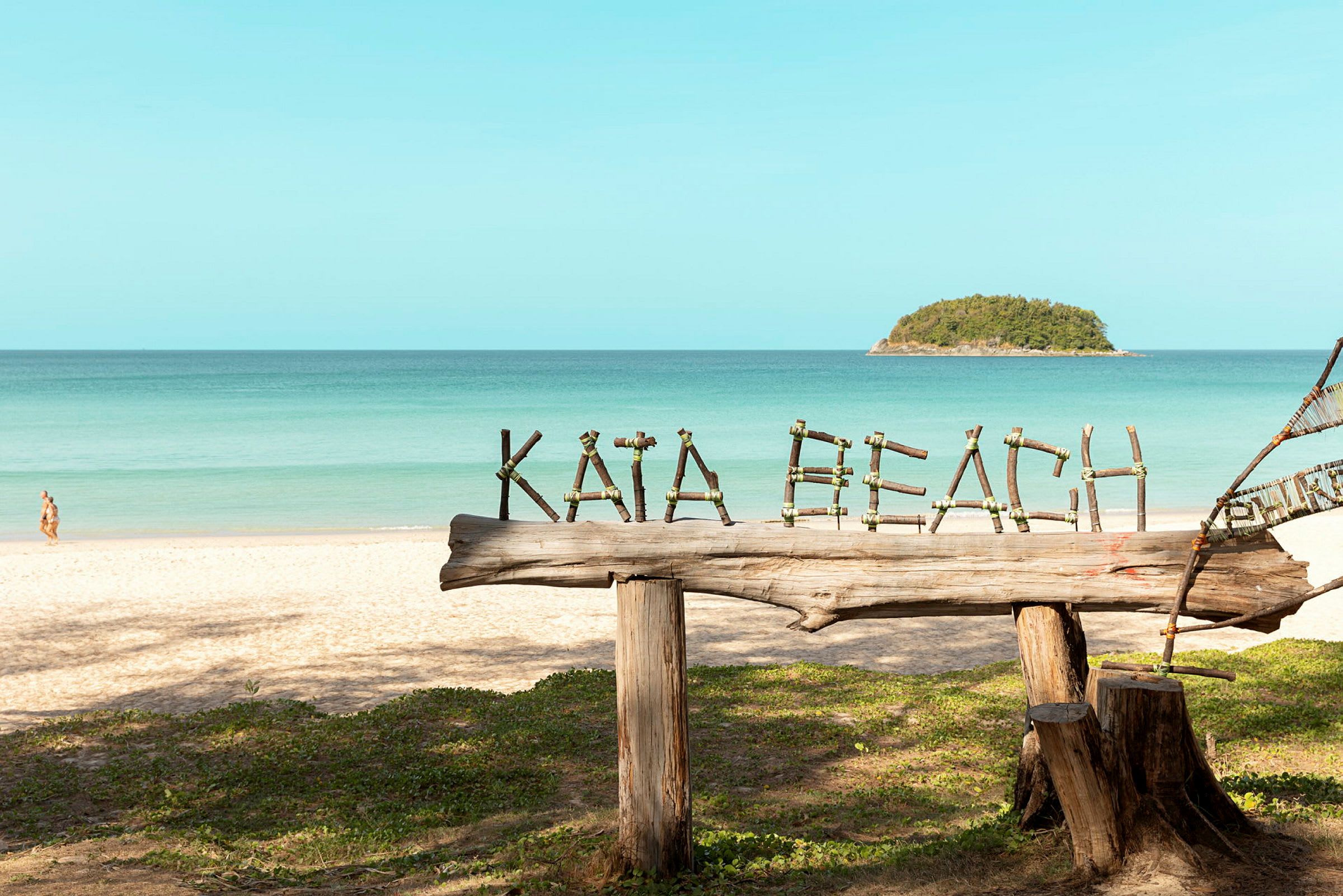 Träskylt med texten 'Kata Beach' på en sandstrand med havet i bakgrunden. (AI-genererad text)