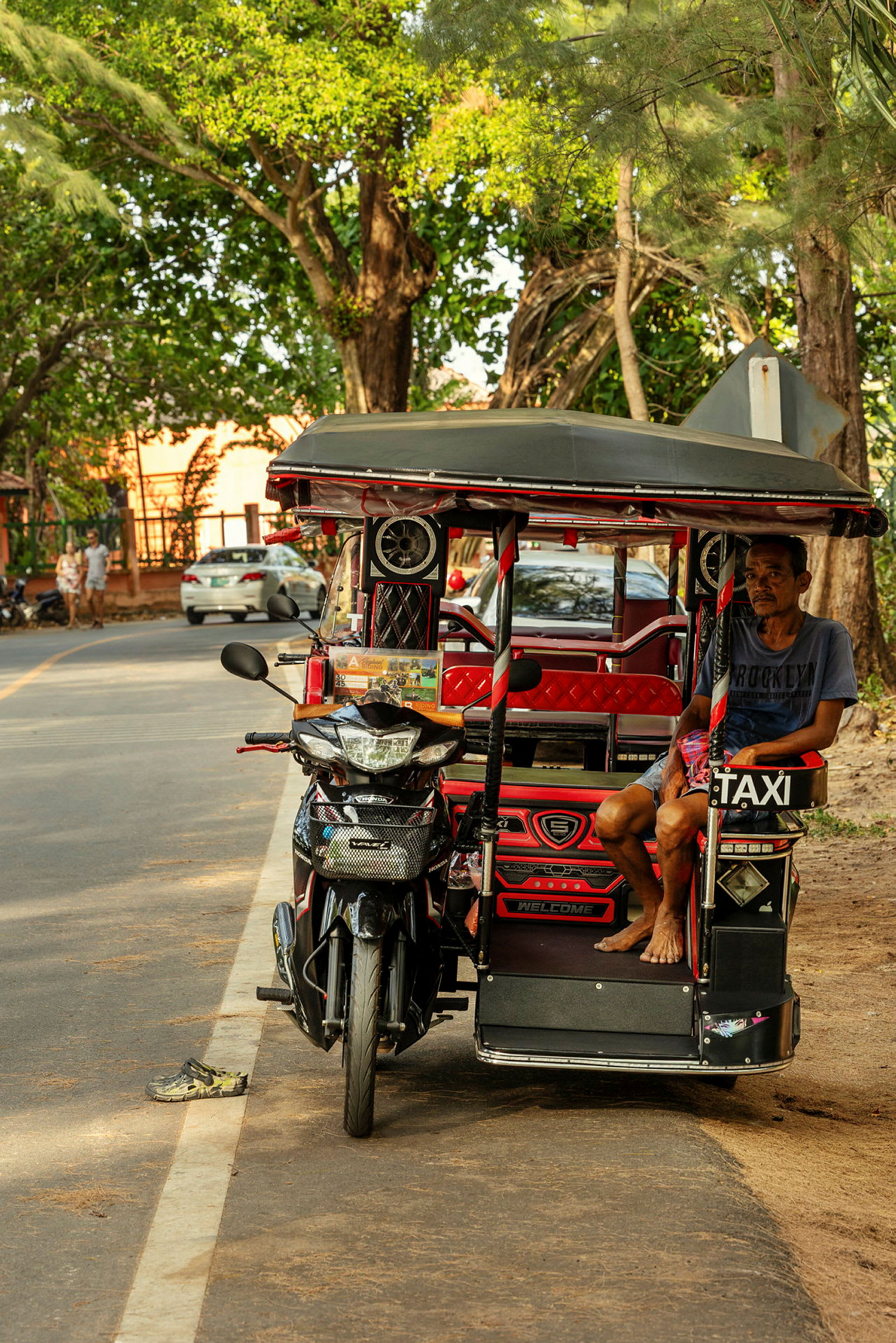 En tuk-tuk står parkerad vid sidan av en gata. En man sitter i fordonet och omgivningen består av träd och några parkerade bilar. (AI-genererad text)