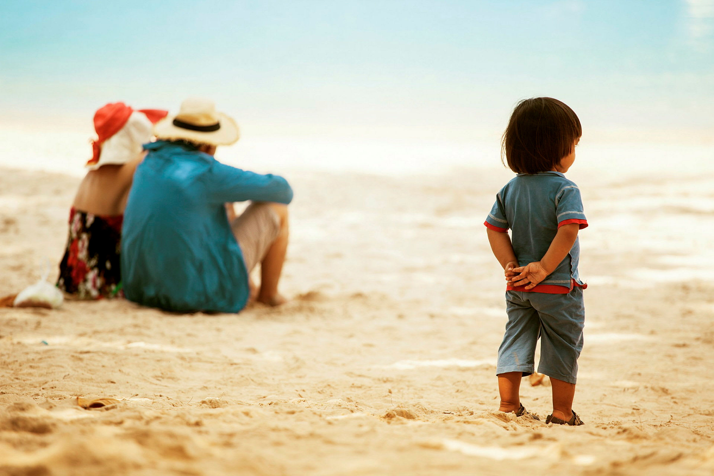 Ett barn står vid stranden med händerna bakom ryggen, medan två vuxna sitter längre bort. (AI-genererad text)