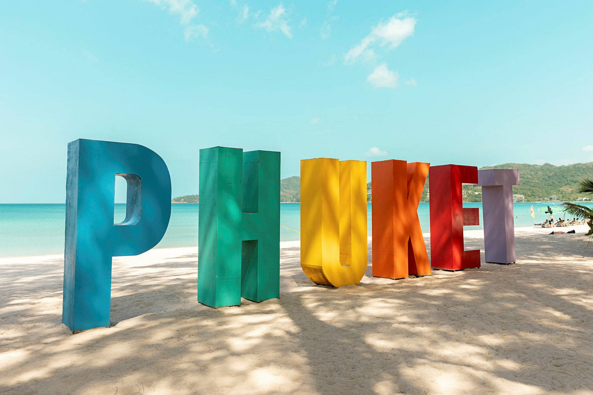 Färgglada bokstäver formar ordet 'Phuket' på en sandstrand, med hav och himmel i bakgrunden. (AI-genererad text)