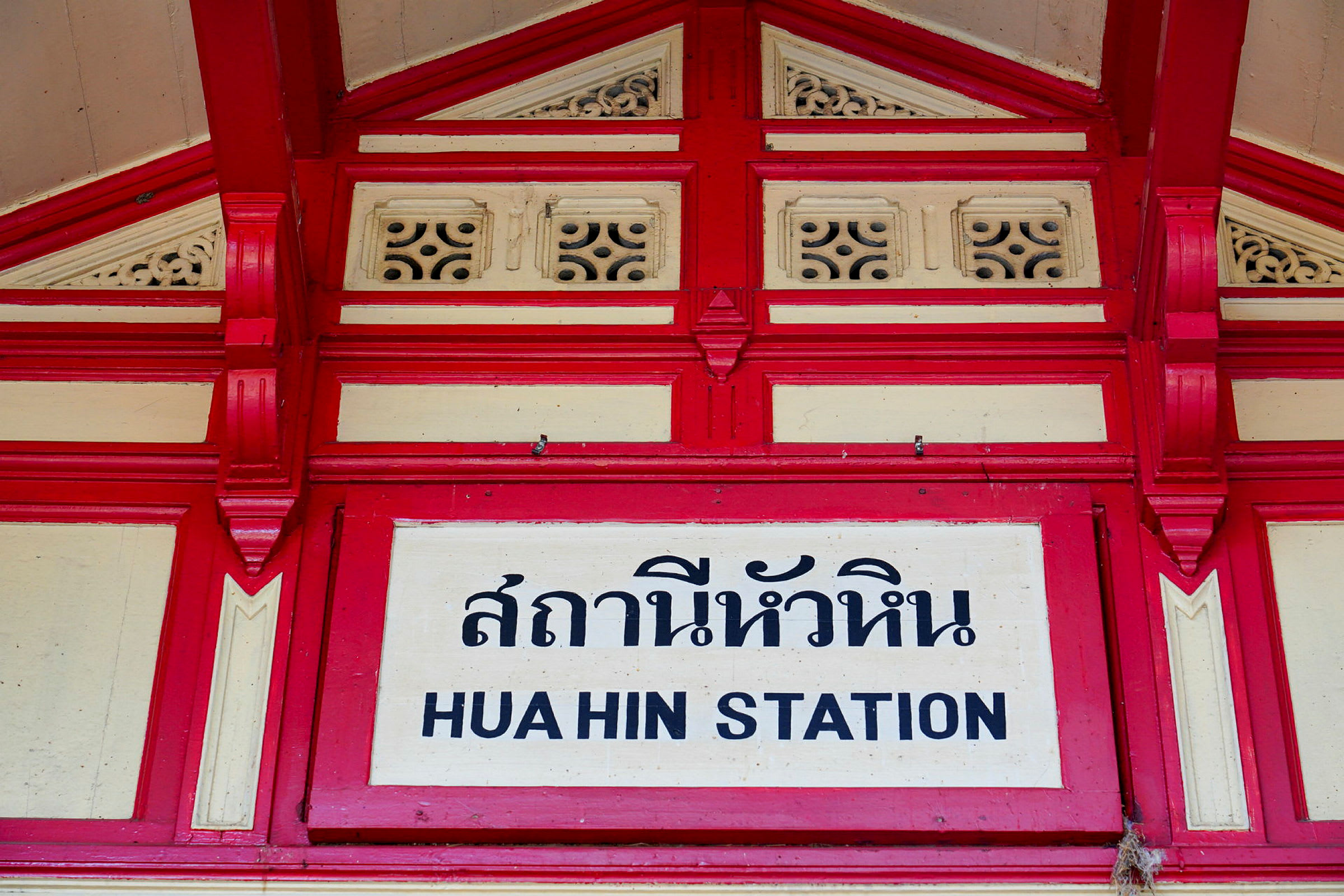 Skylt med texten 'Hua Hin station' med thailändska tecken och dekorativ röd och vit ram. (AI-genererad text)