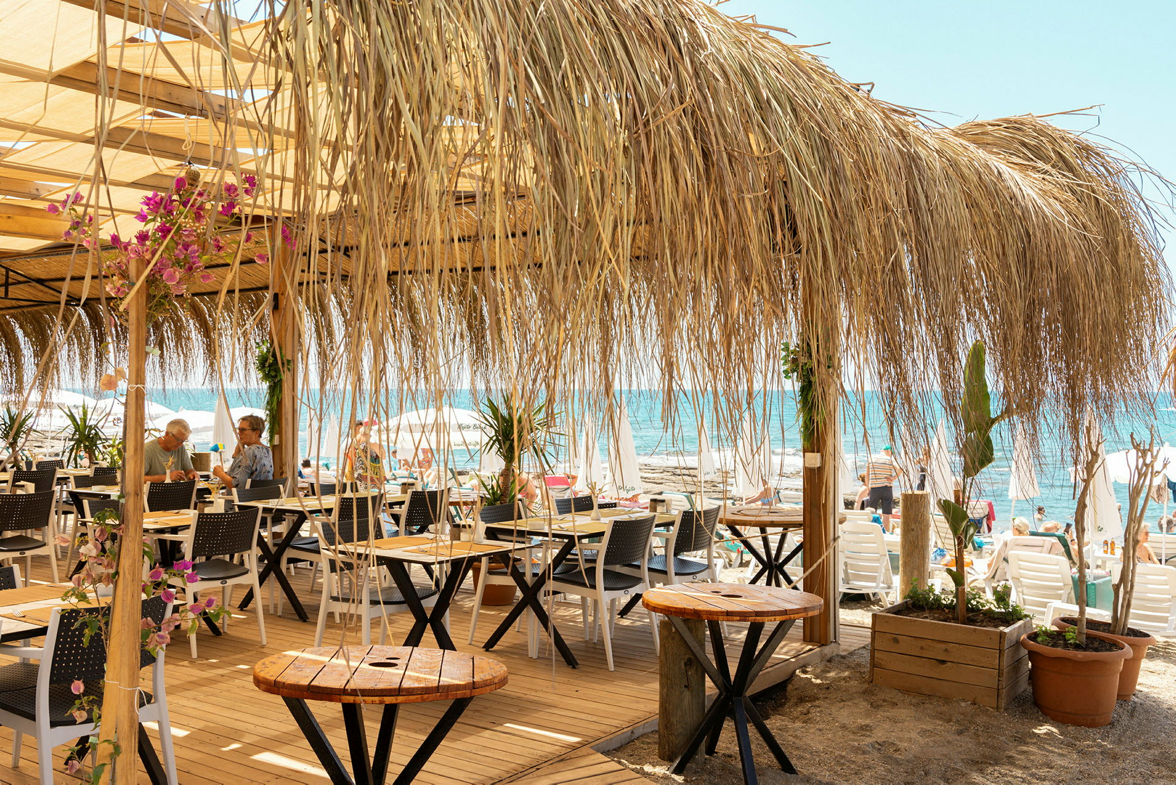 Strandnära café med bord, stolar och halmtak, med utsikt över havet. (AI-genererad text)