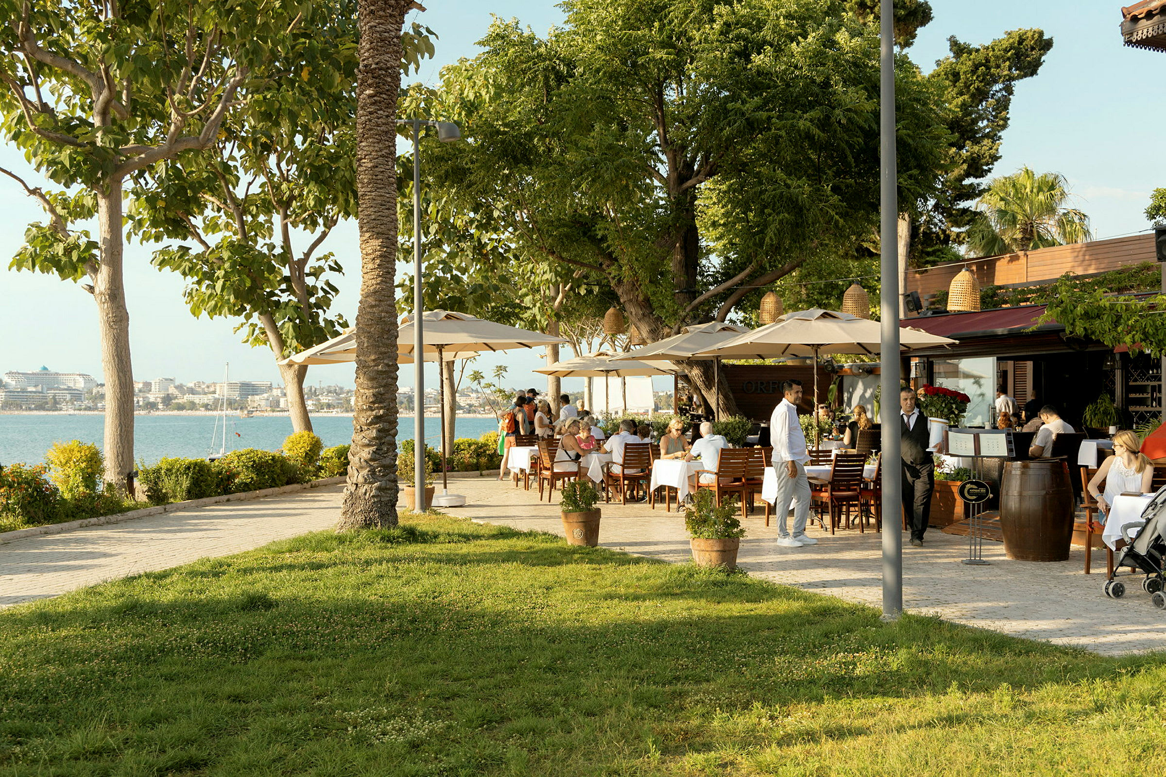 Restaurang med uteservering, bord med vita dukar och grönskande träd nära vattnet. (AI-genererad text)