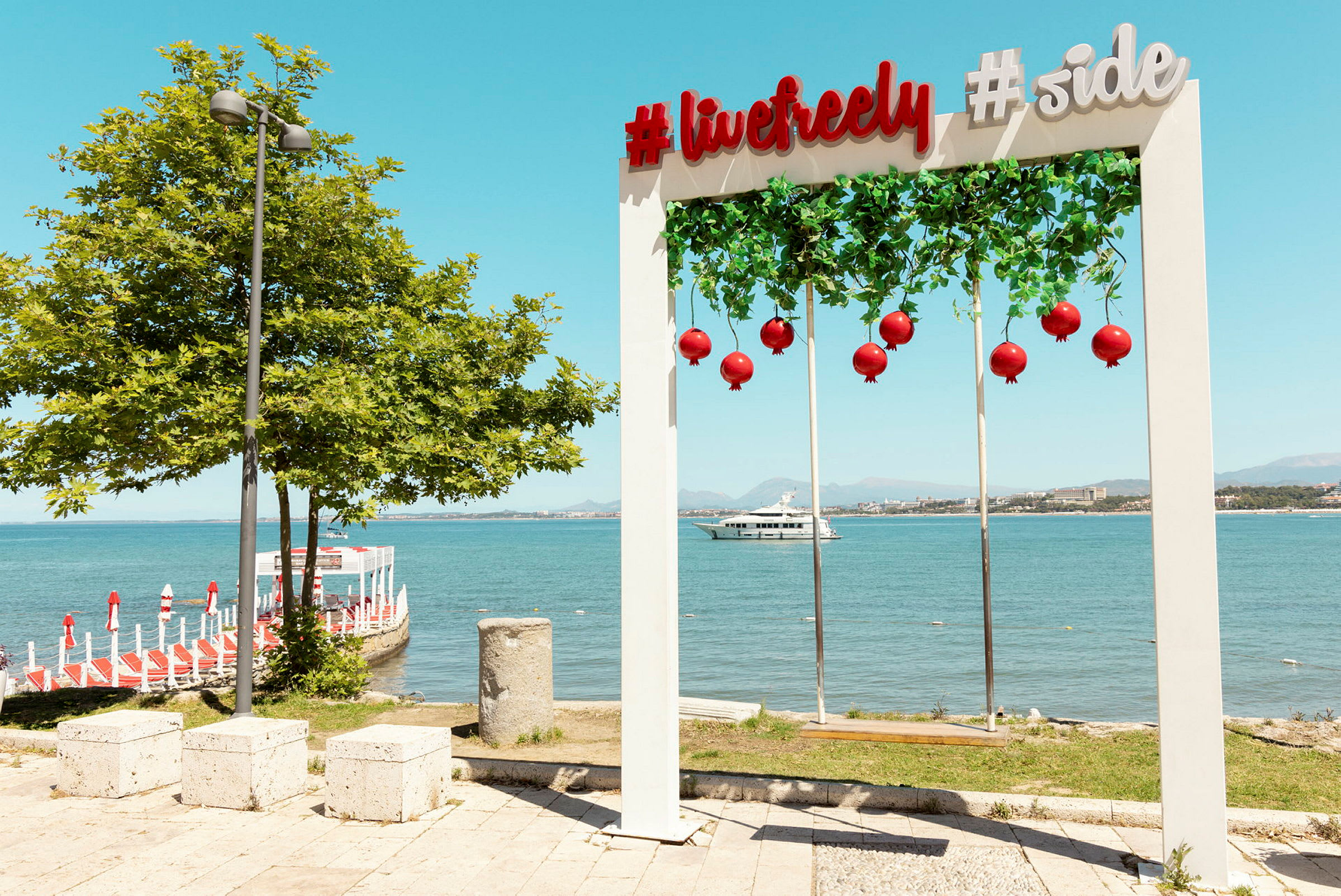 En dekorativ båge med texten '#livefreely #side', hängande röda dekorationer, vid havet framför en brygga och en yatch i bakgrunden. (AI-genererad text)