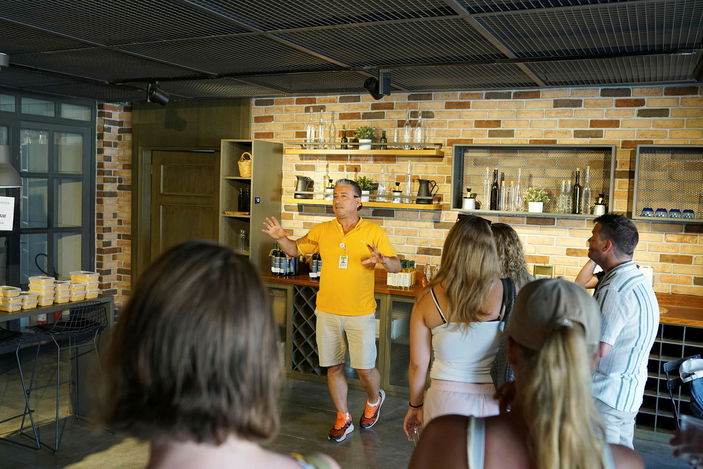 En person i gul tröja håller en presentation framför en grupp människor i ett rum med tegelväggar och köksinredning. (AI-genererad text)