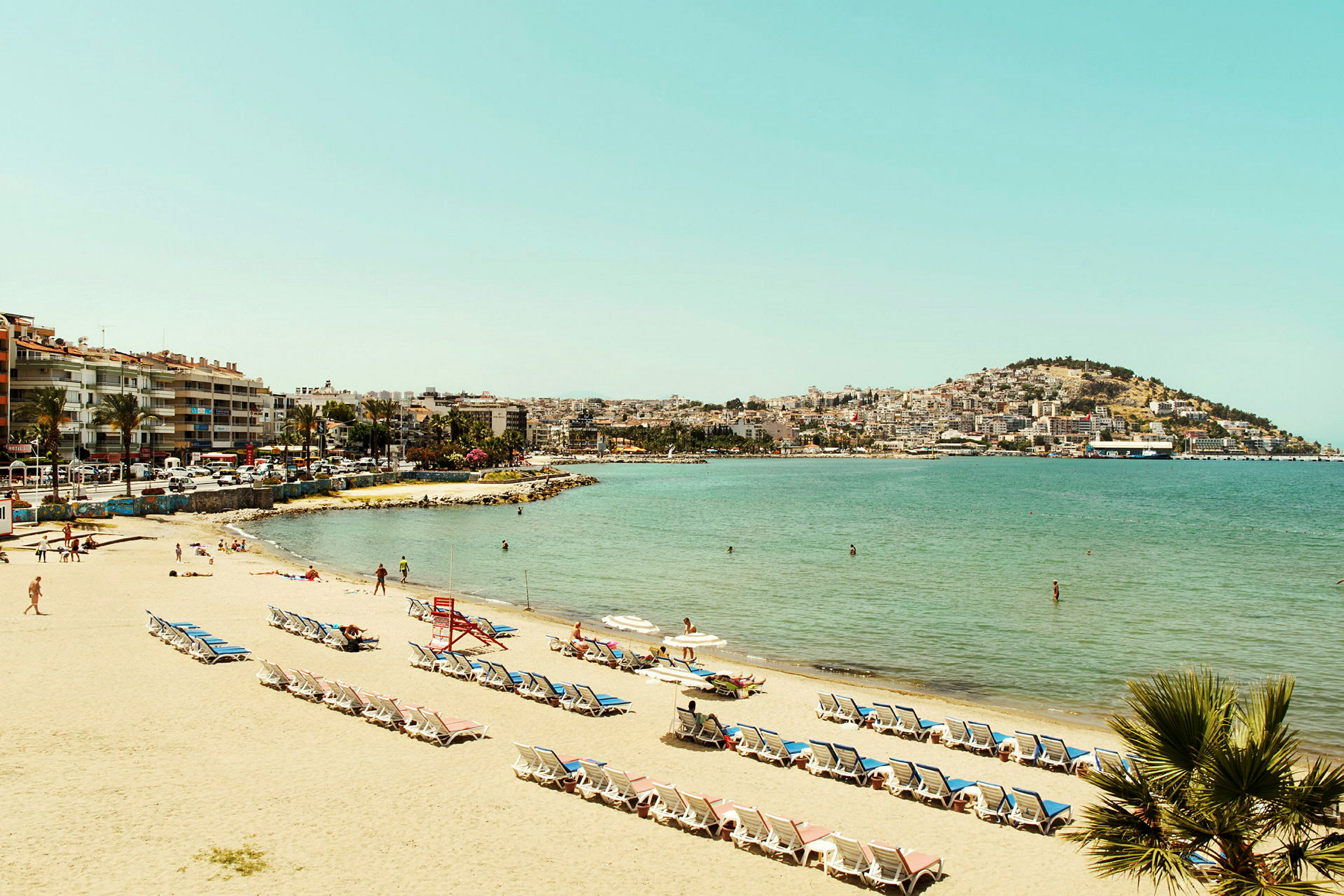 Strand med solstolar och parasoller, människor vid vattnet, med en stad i bakgrunden. (AI-genererad text)
