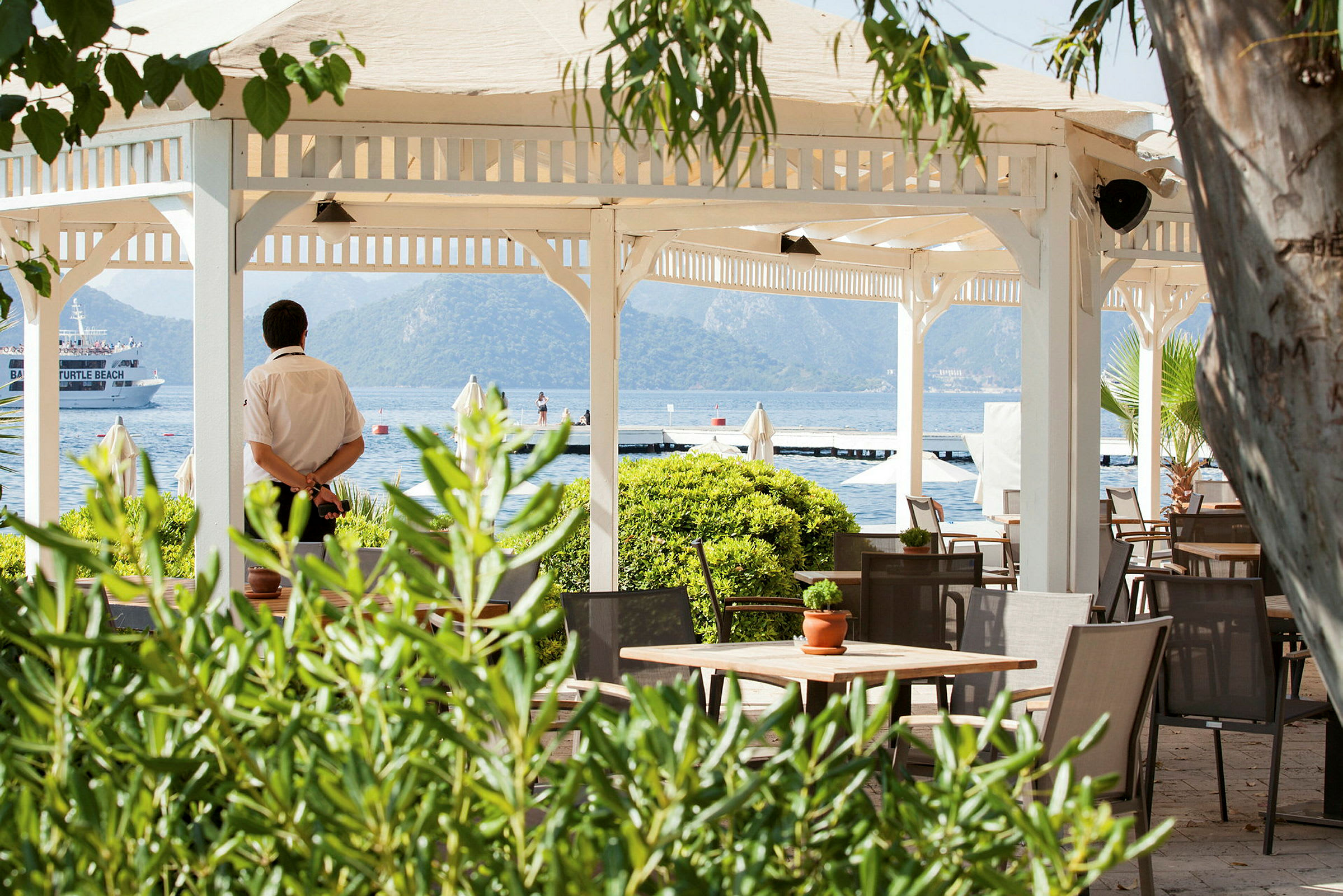 En öppen paviljong med bord och stolar vid havet, omgiven av grönska. (AI-genererad text)