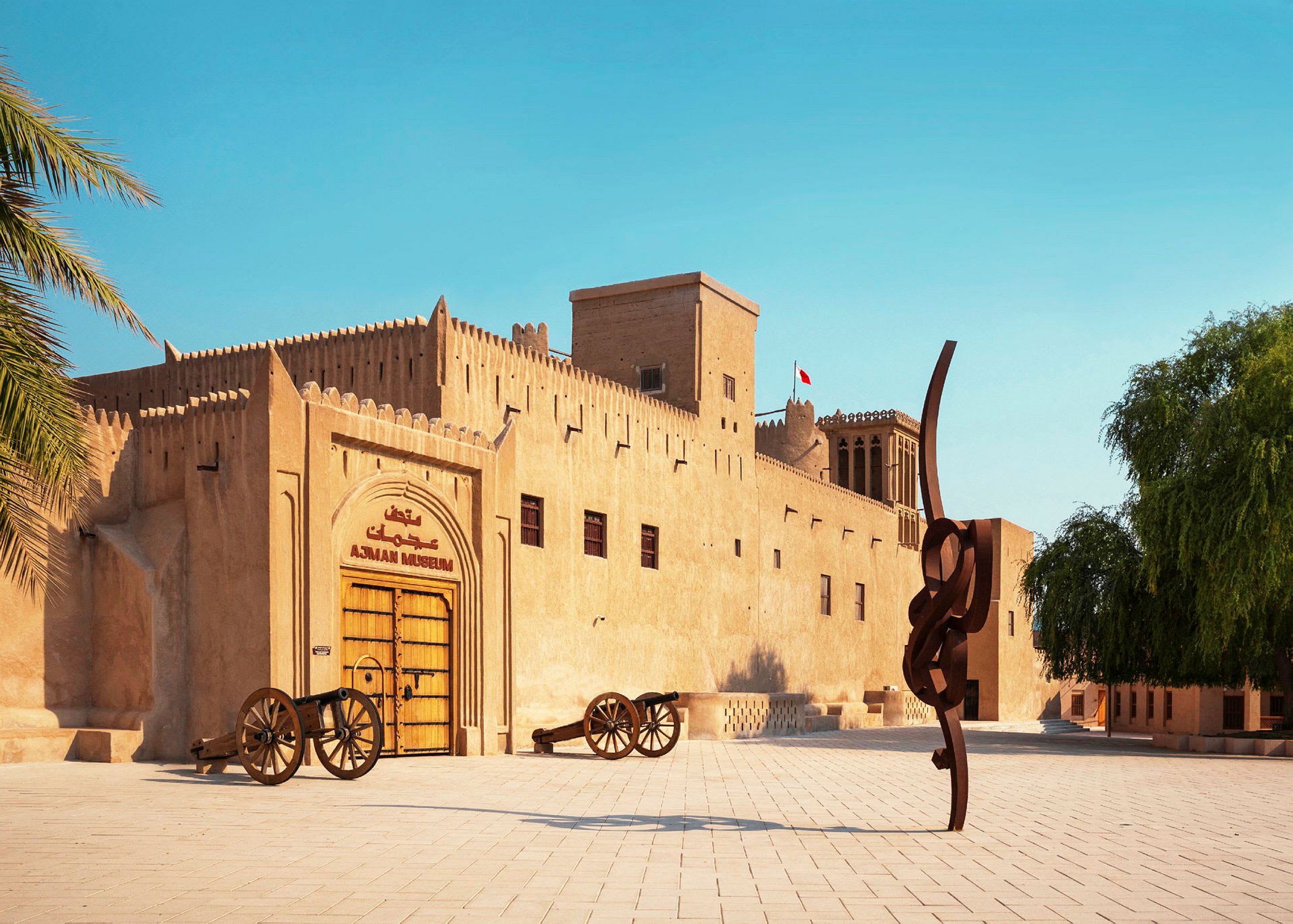 En historisk byggnad med sandfärgade fasader, det står "Ajman Museum" ovanför en stor trädörr. Framför byggnaden finns två kanoner och en modern skulptur. (AI-genererad text)