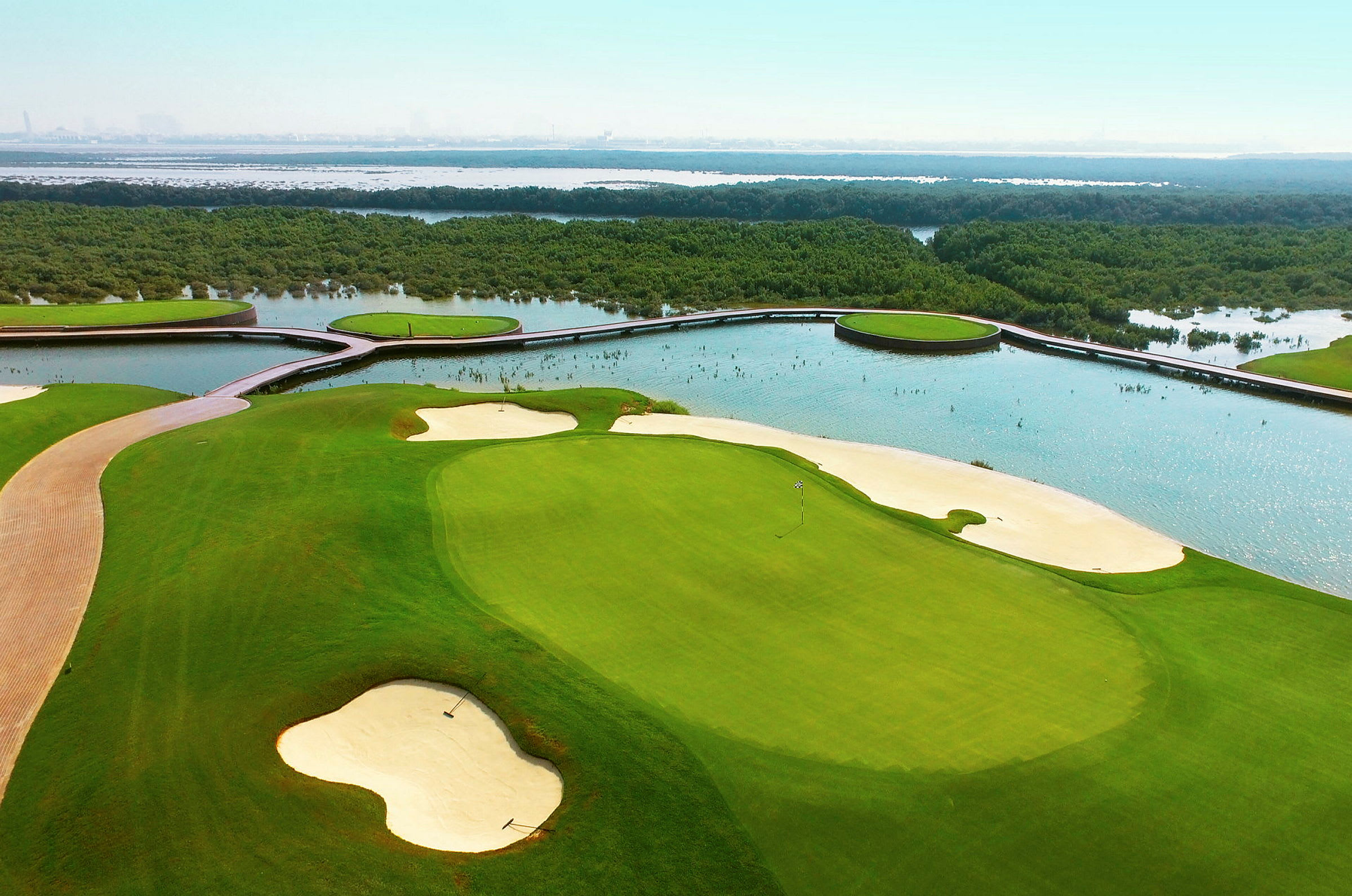 Bilden visar en golfbana med sandbunkrar och gröna fairways, omgiven av vatten och vegetation. (AI-genererad text)