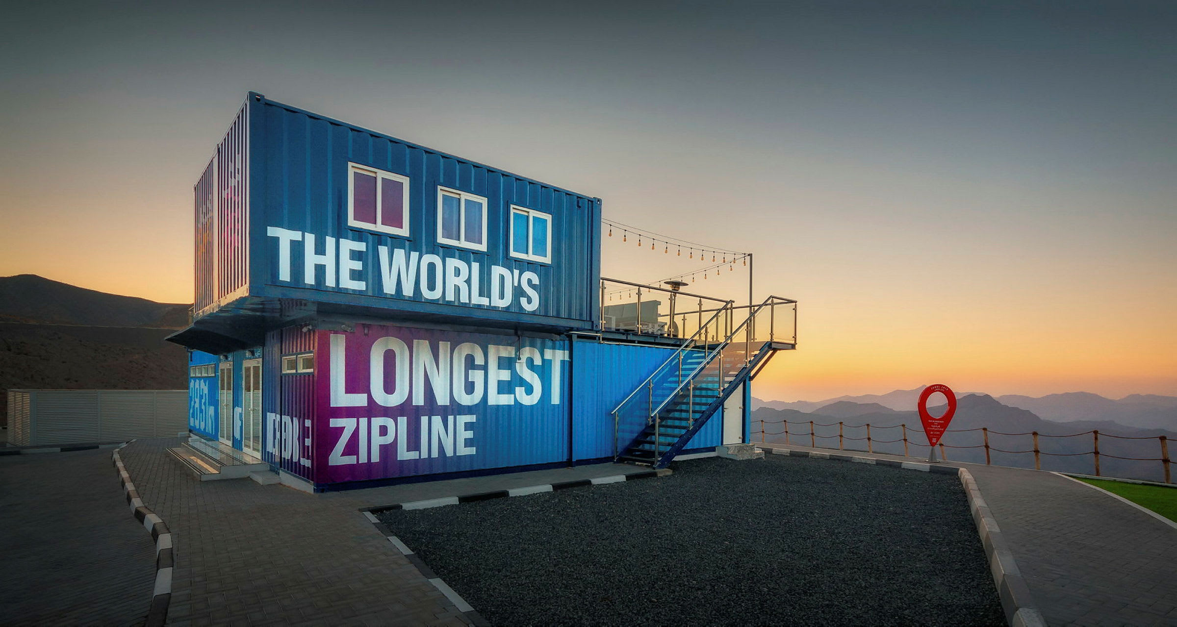 Blå och lila byggnad med texten 'The World's Longest Zipline' vid solnedgång, omgiven av berg. (AI-genererad text)