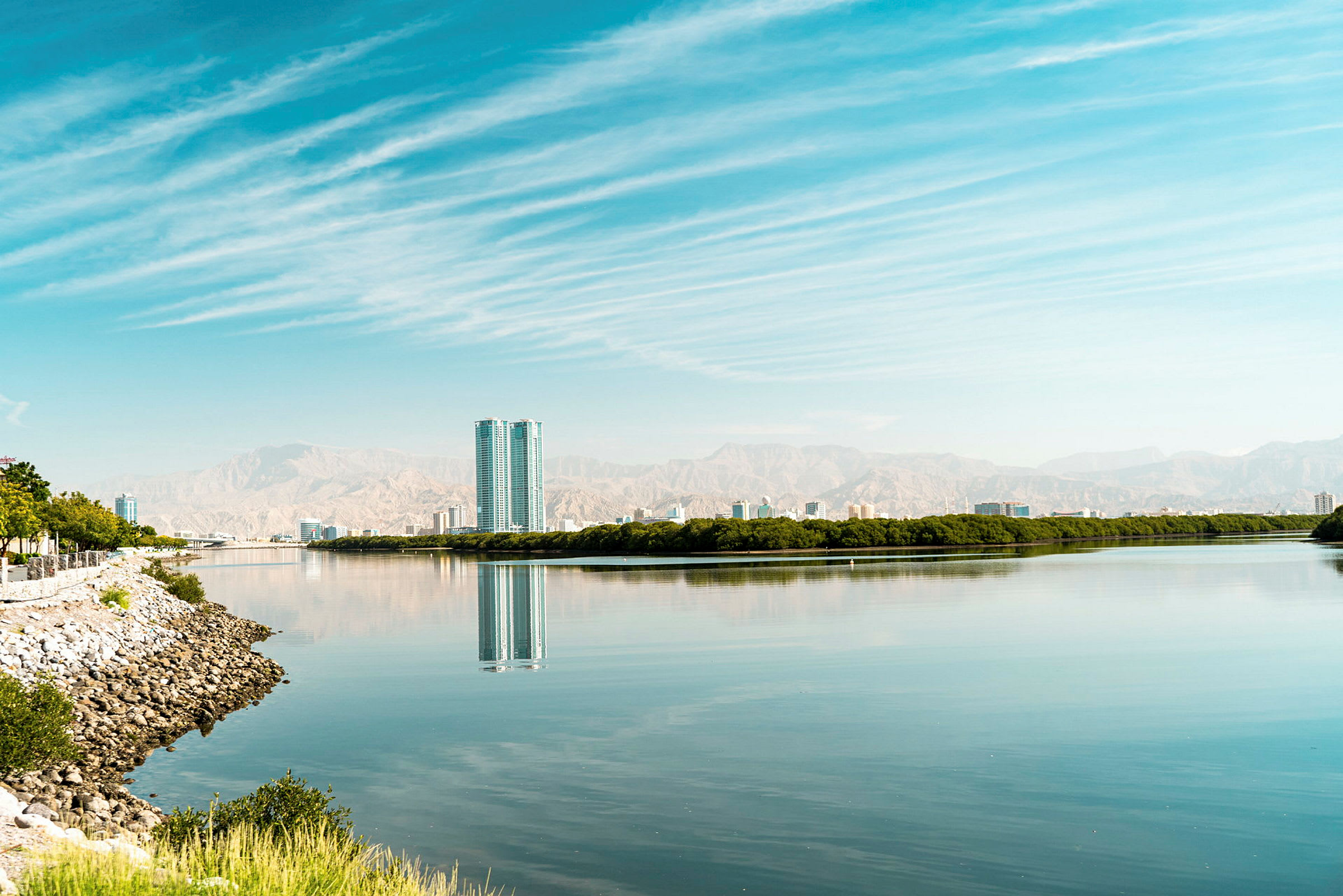 En lugn vattenyta reflekterar en stadssiluett och omgivande grönska, med berg i bakgrunden. (AI-genererad text)