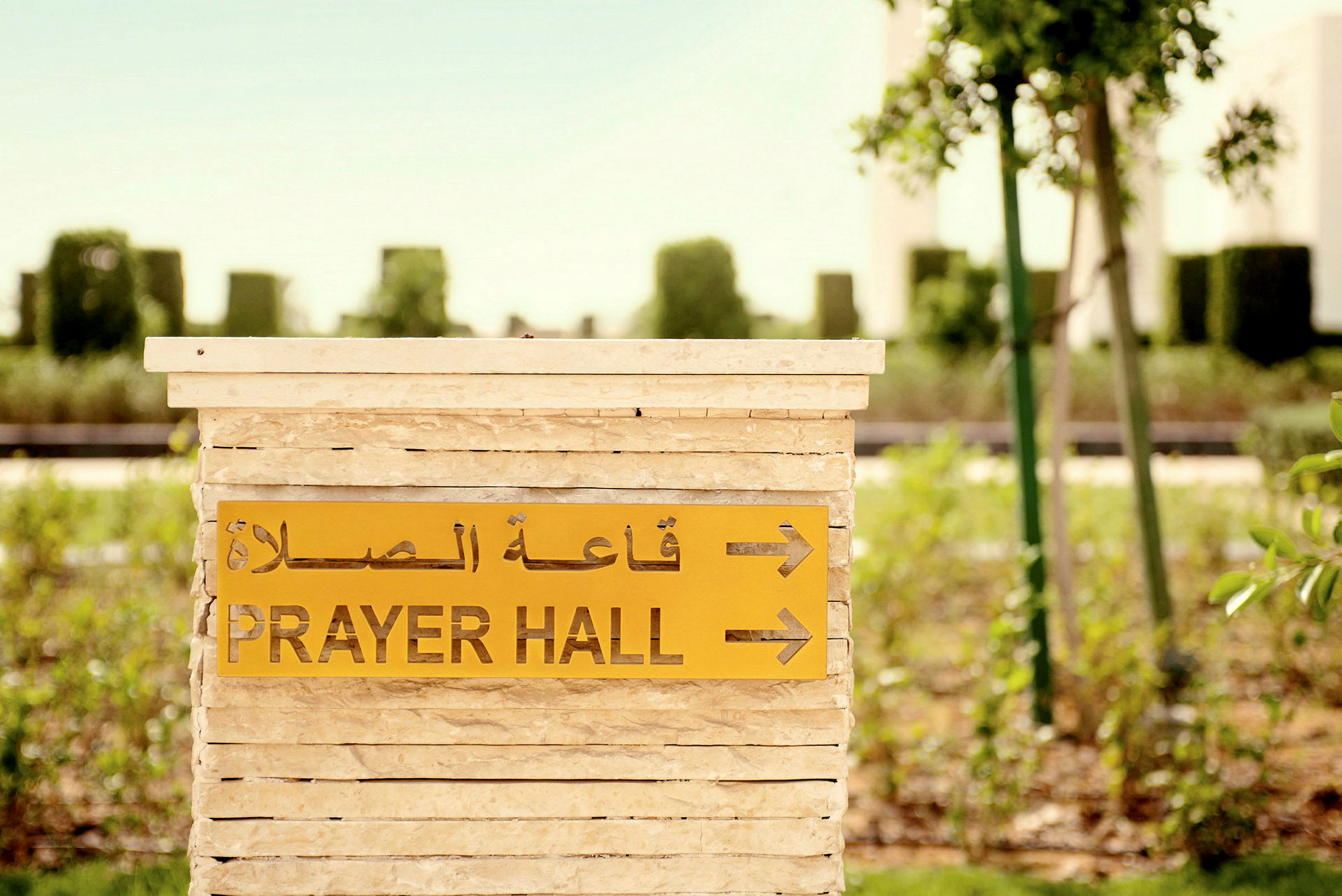 En skylt med texten 'Prayer Hall' och arabisk text pekar mot en bönehall, omgiven av grönska. (AI-genererad text)