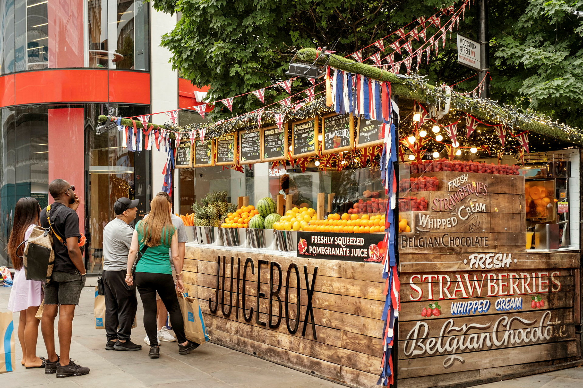 En frukt- och juicekiosk med dekorativa flaggor och skyltar. (AI-genererad text)