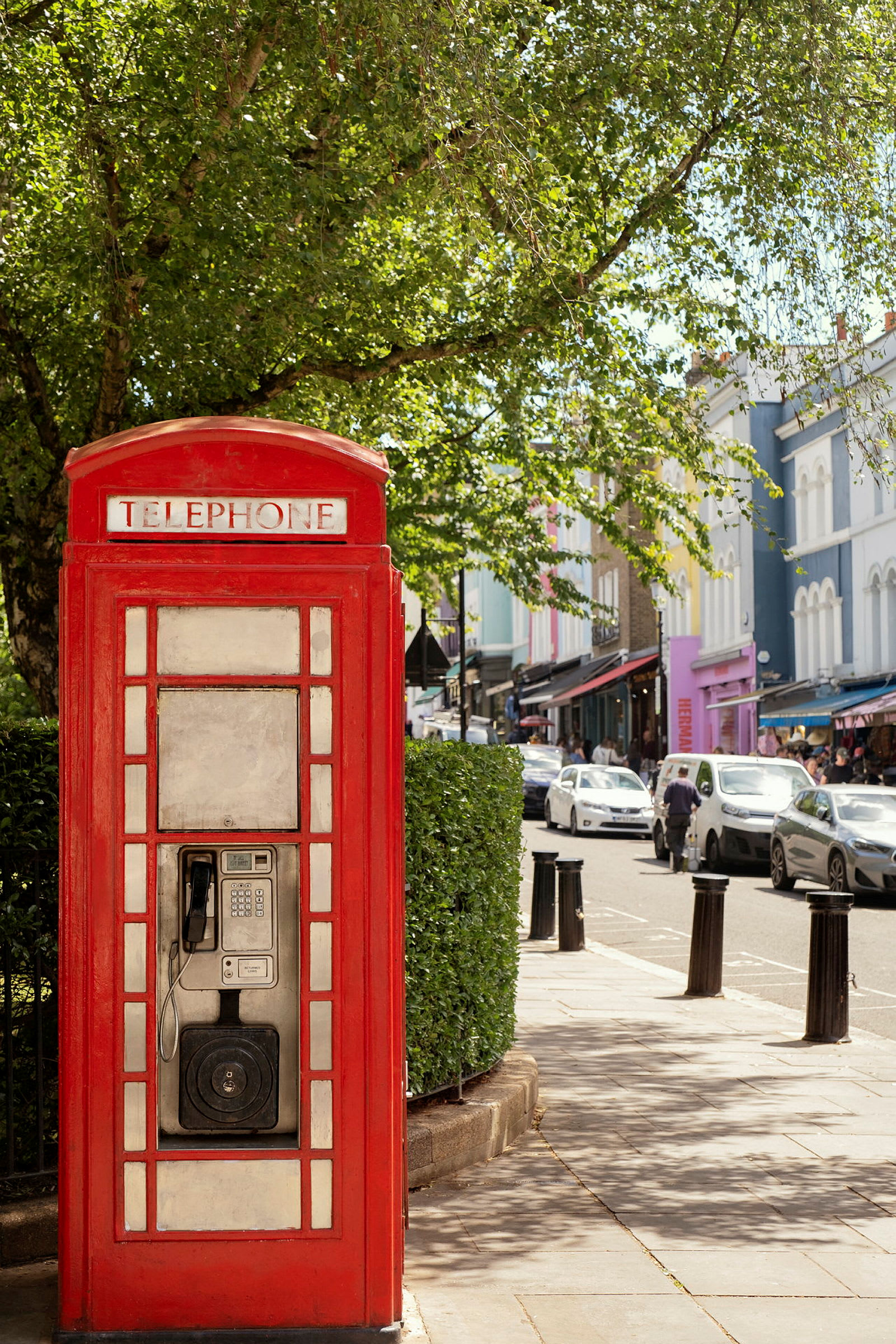 Röd telefonkiosk med grönska och stadsgata i bakgrunden. (AI-genererad text)