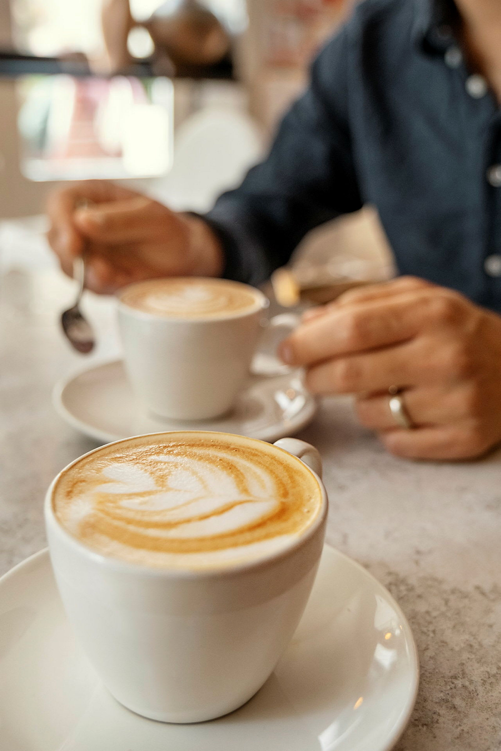 Två cappuccino i vita koppar på ett bord, med en hand som håller i en sked. (AI-genererad text)