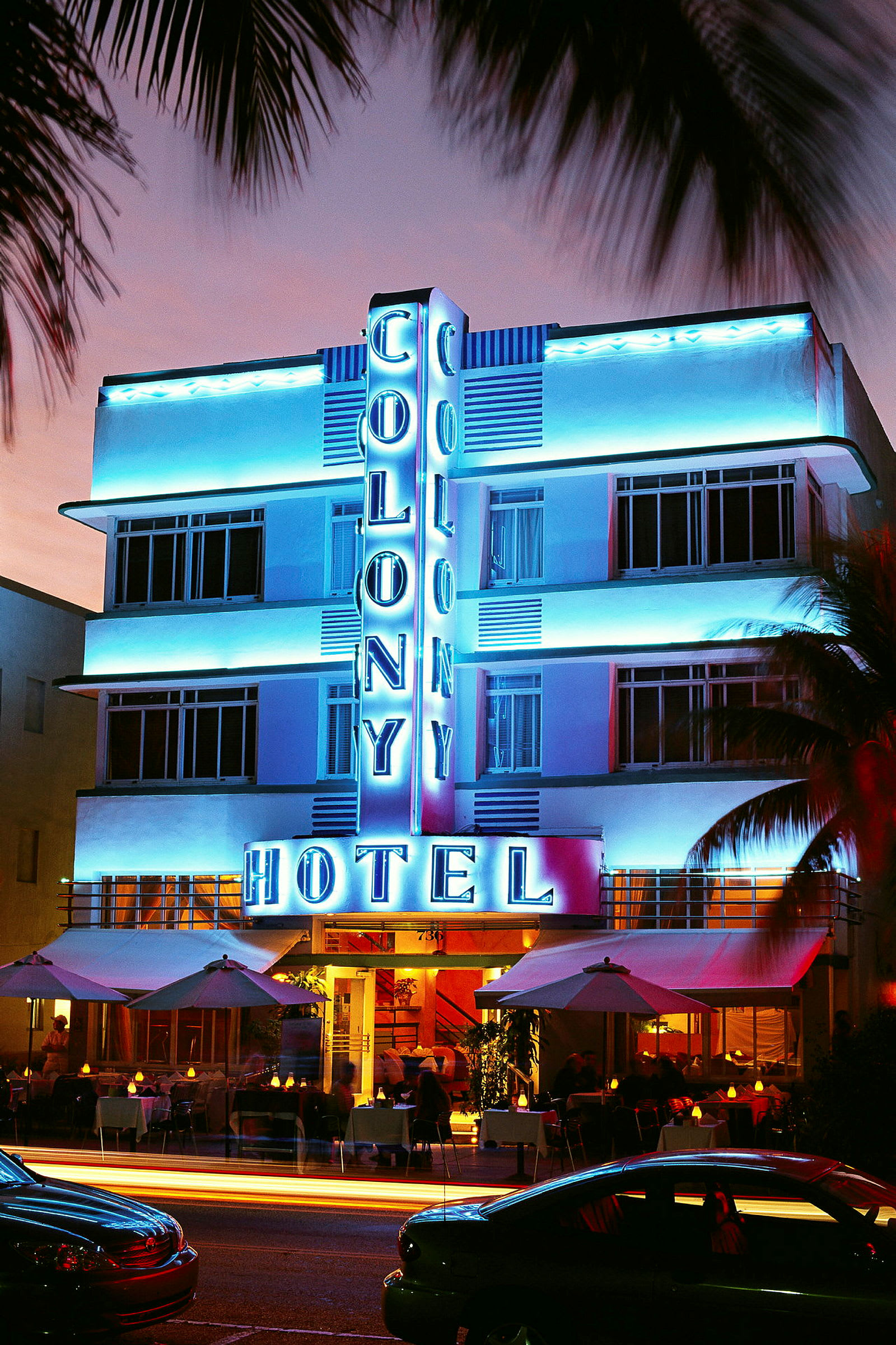 Exteriör av Colony Hotel upplyst i neon om natten med utomhusservering och fordon framför. (AI-genererad text)