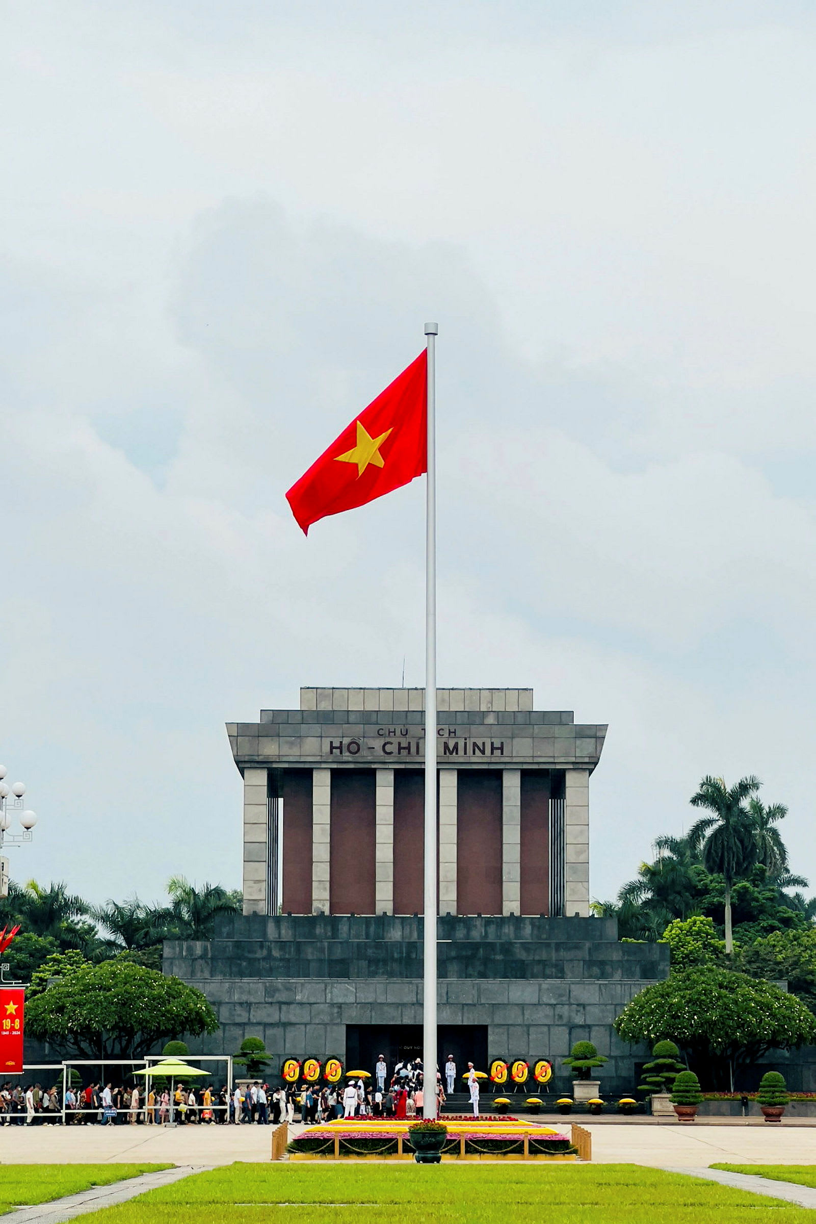 Ett stort mausoleum med texten 'Ho Chi Minh' på fasaden, framför vilket den vietnamesiska flaggan står uppsatt på en flaggstång. Människor samlas framför byggnaden som omges av träd och gräsmattor. (AI-genererad text)