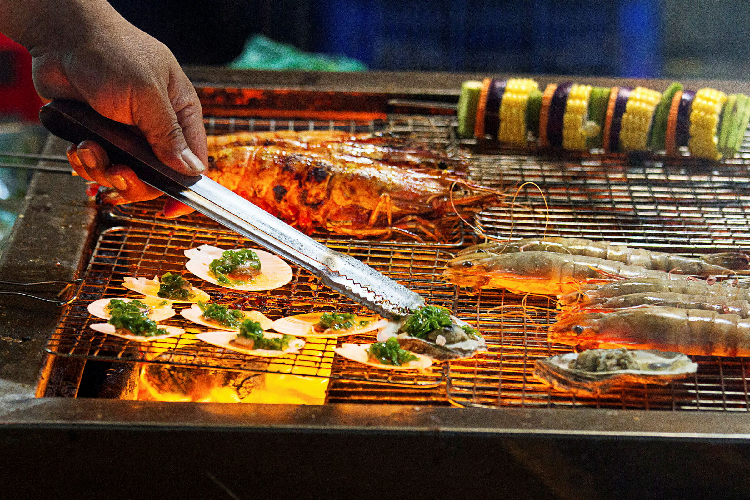 En grill med skaldjur och grönsaksspett, där en hand hanterar maten med tång. (AI-genererad text)