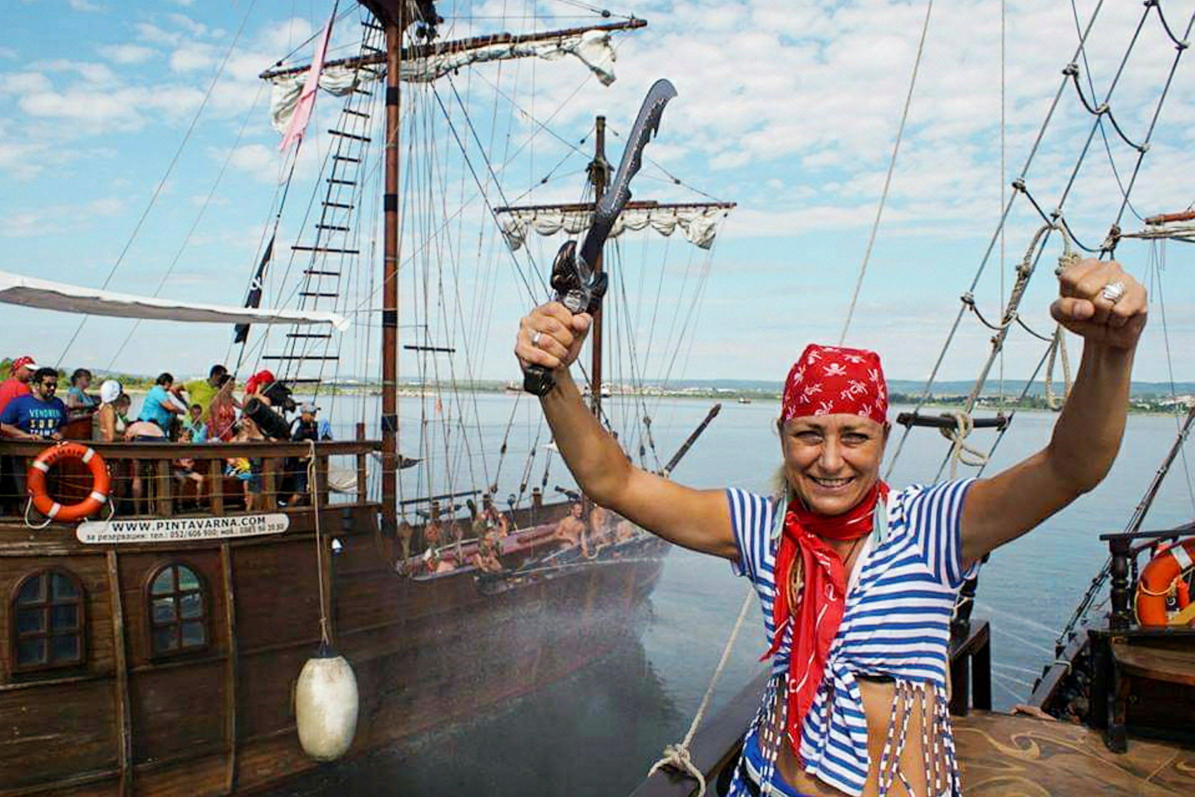 En person klädd som pirat står på ett skepp och håller upp en leksakssabel med människor i bakgrunden. Skeppet är dekorerat och utrustat som ett gammaldags segelfartyg. (AI-genererad text)