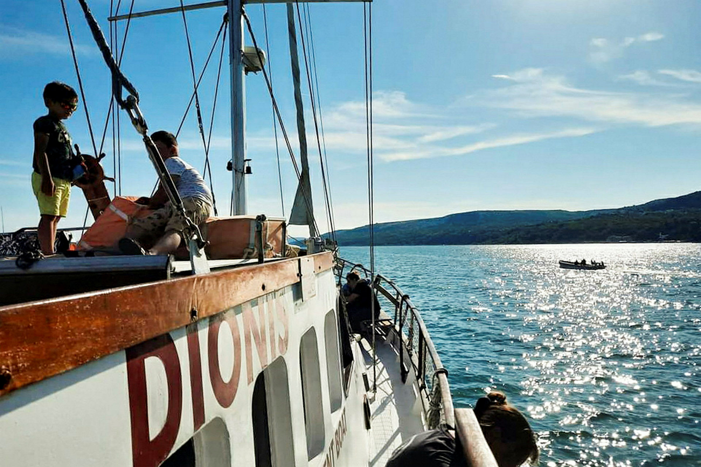 En segelbåt på en solig dag med människor ombord, och en mindre båt syns i bakgrunden på det glittrande vattnet. (AI-genererad text)