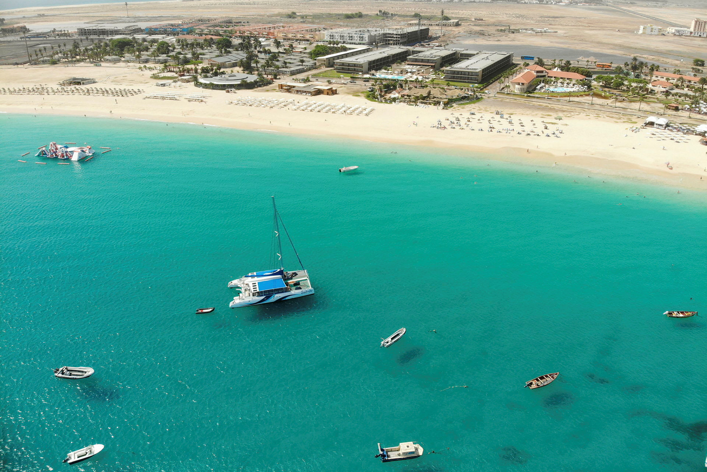 Luftvy över strand med vit sand och turkost hav. Flera båtar flyter nära stranden och byggnader syns i bakgrunden. (AI-genererad text)